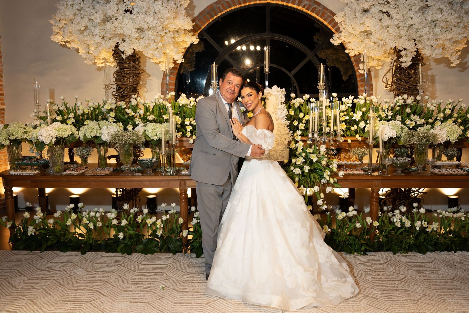 Bride and groom pose elegantly in front of a stunning floral wedding backdrop with white flowers and candlelight.