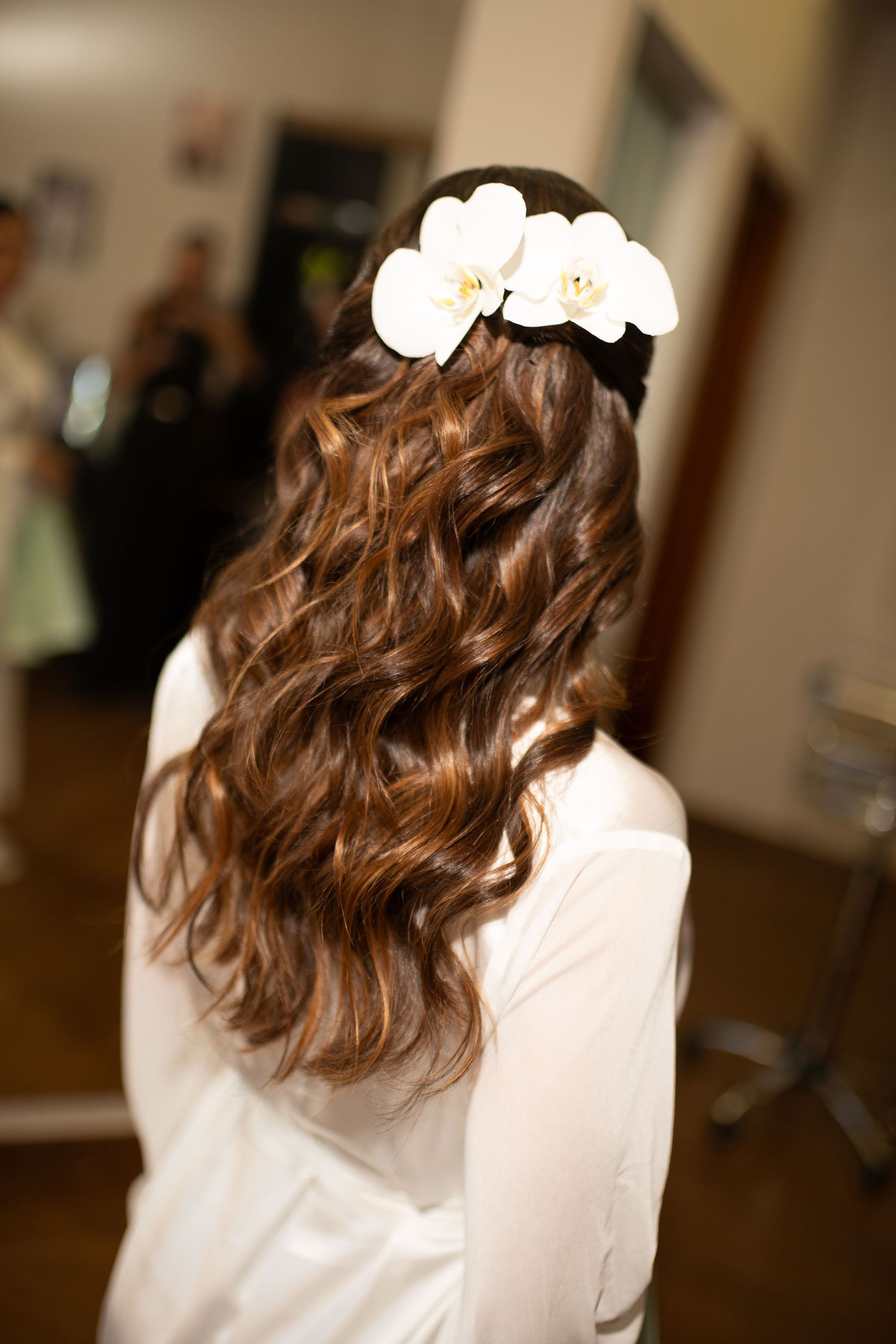 Bride with long curly brown hair adorned with white orchid flowers, viewed from behind in a white robe.