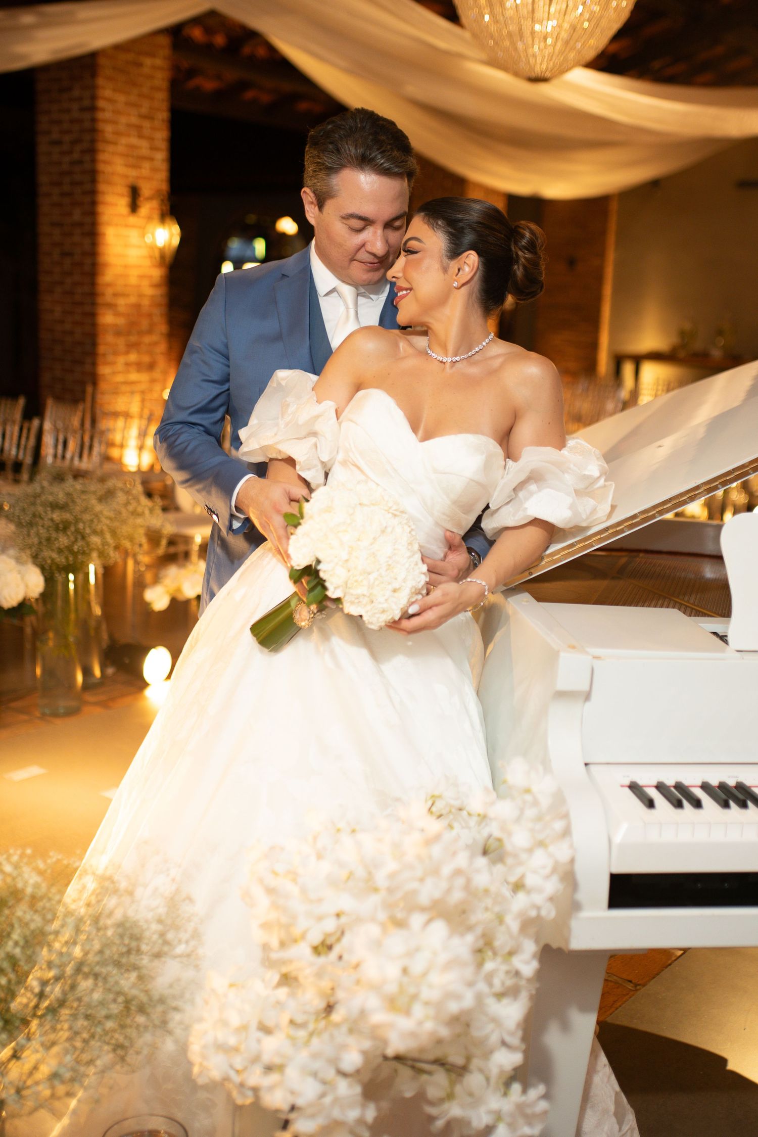 Bride in white off-shoulder gown holds bouquet beside groom in blue suit near white grand piano at elegant wedding reception.