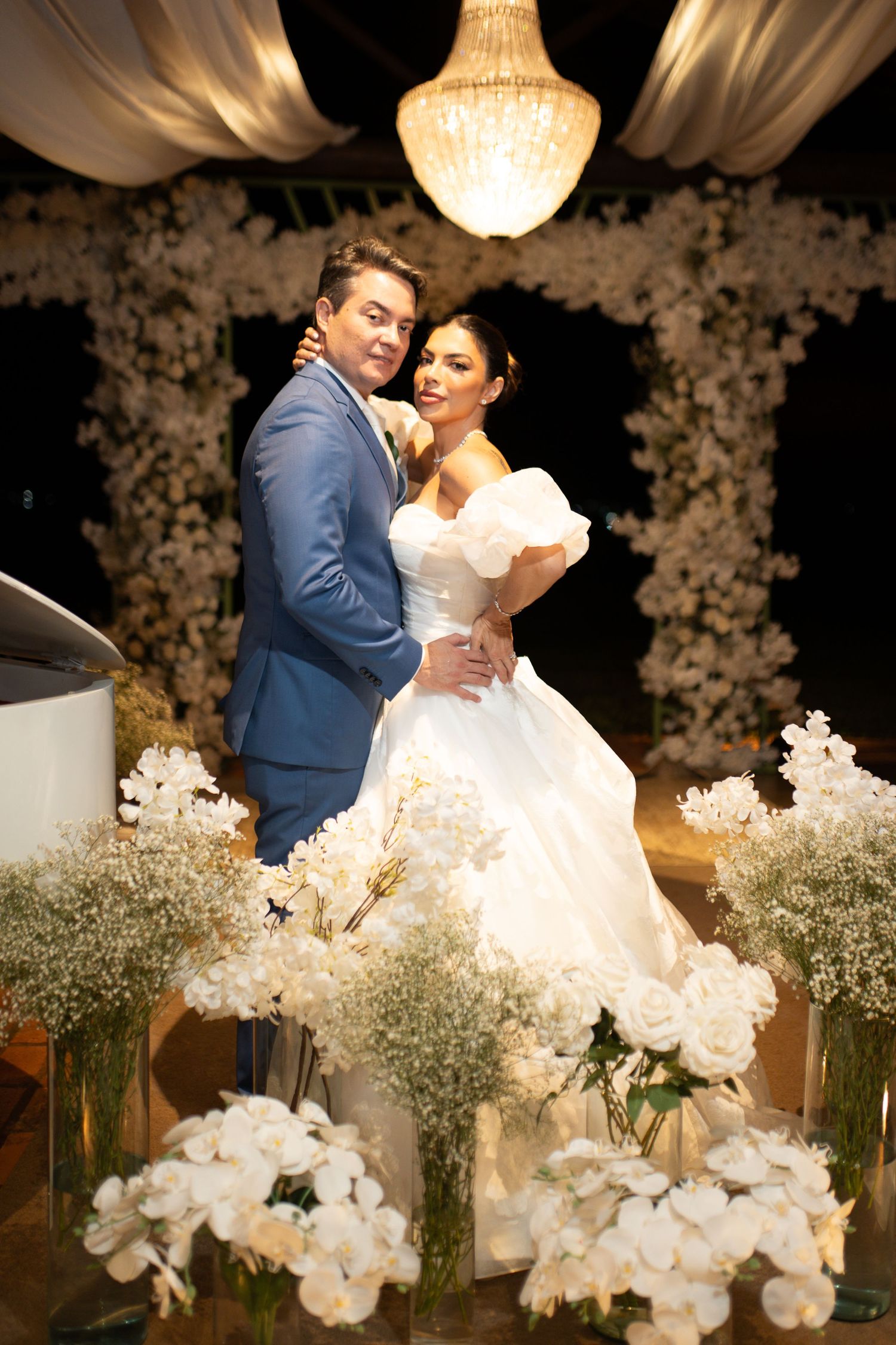 Bride in white off-shoulder gown and groom in blue suit embrace at elegant wedding ceremony with white floral arrangements.