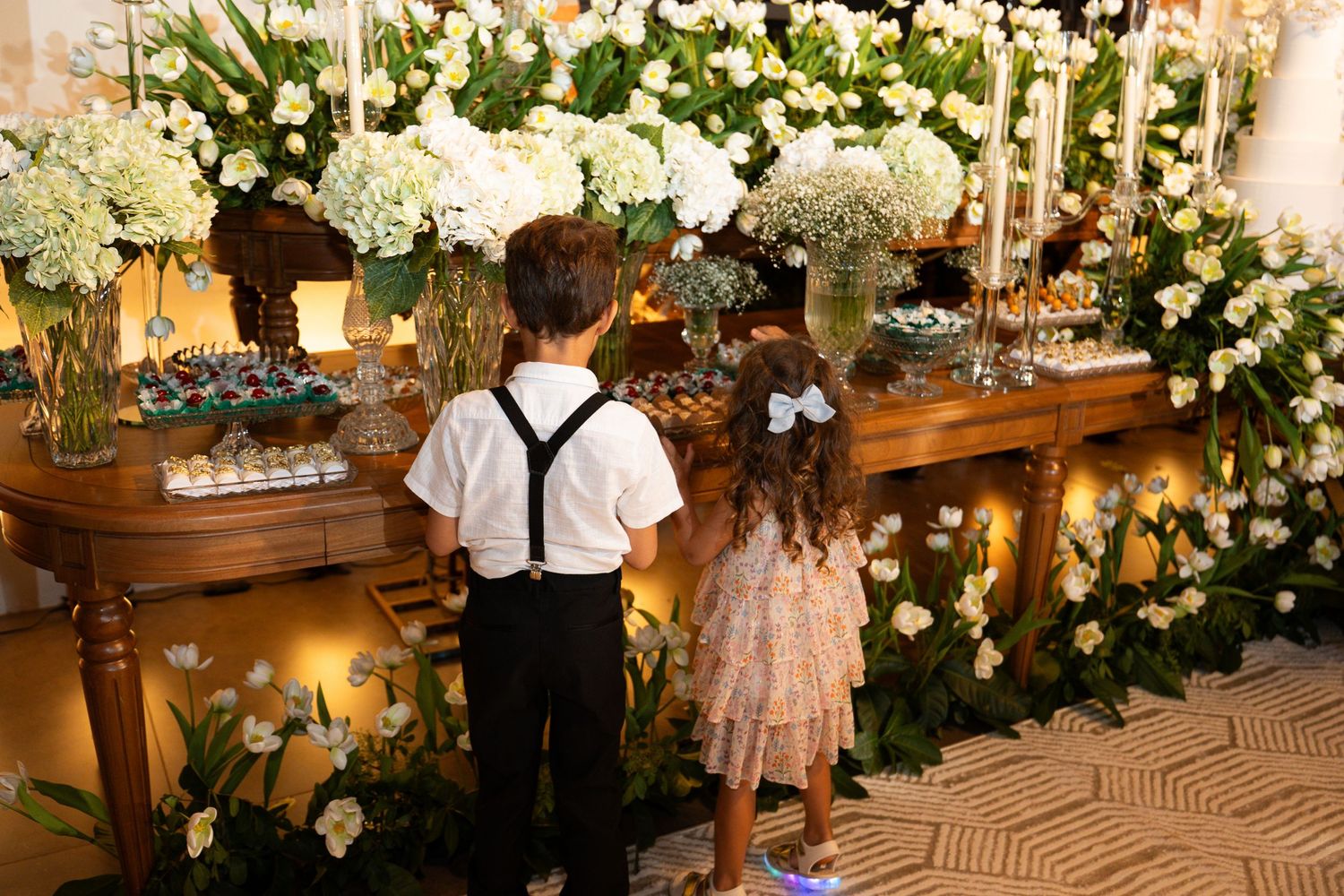 Two children in formal attire stand before an elegant floral display with white flowers at a wedding reception.