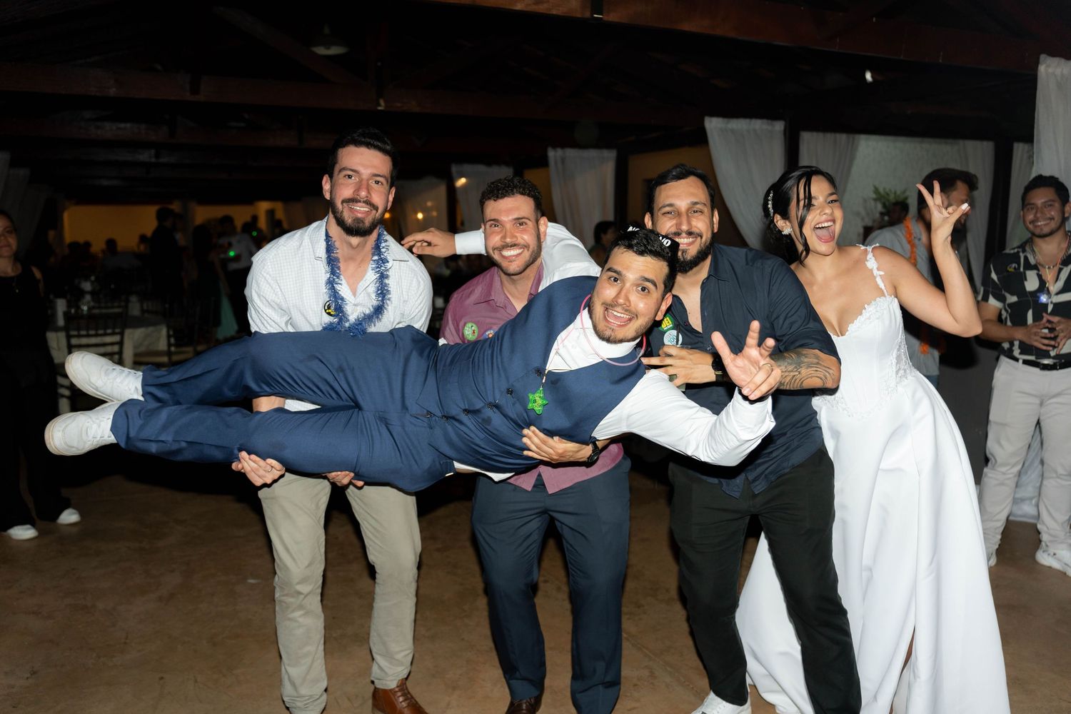 Groomsmen lifting groom horizontally at wedding reception while bride poses excitedly beside them.