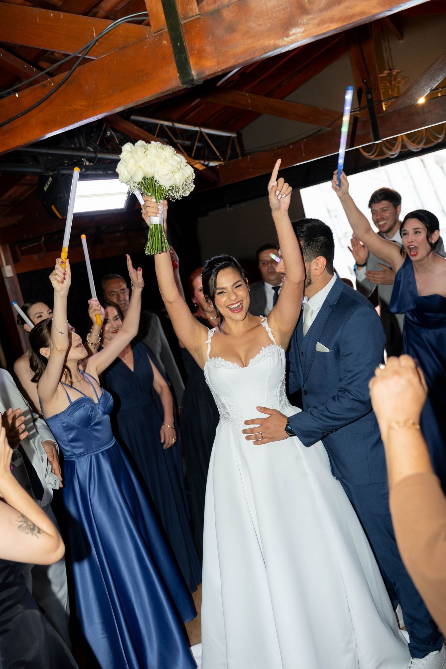 Joyful bride in white gown raises bouquet surrounded by wedding party in navy blue attire celebrating at indoor reception.
