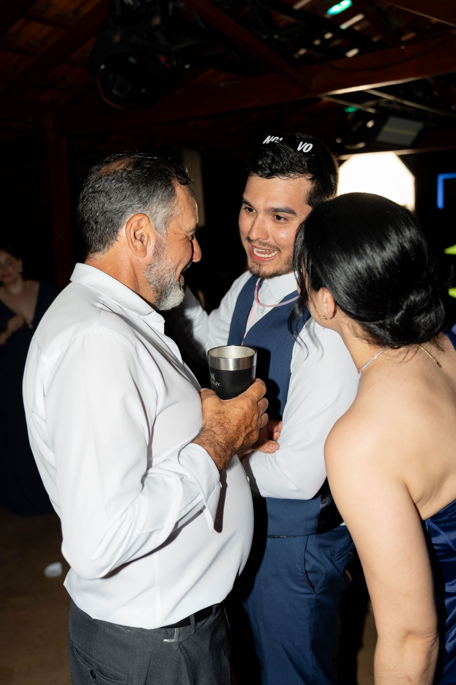 Groom in blue vest shares a joyful moment with an older man and woman in elegant attire at a wedding reception.