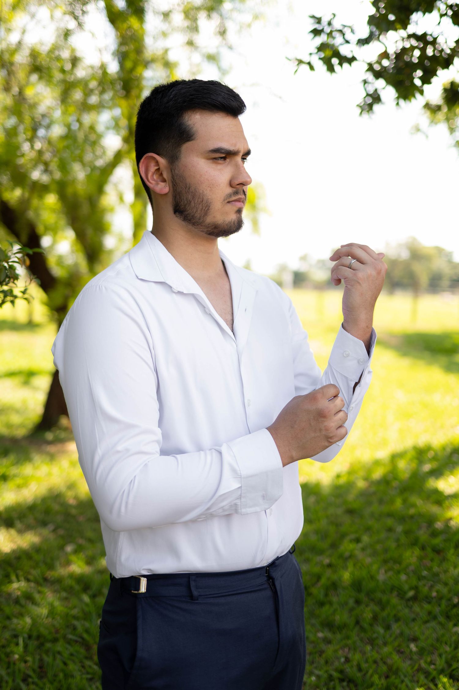Young man in white dress shirt adjusting cufflinks outdoors in a green park setting.