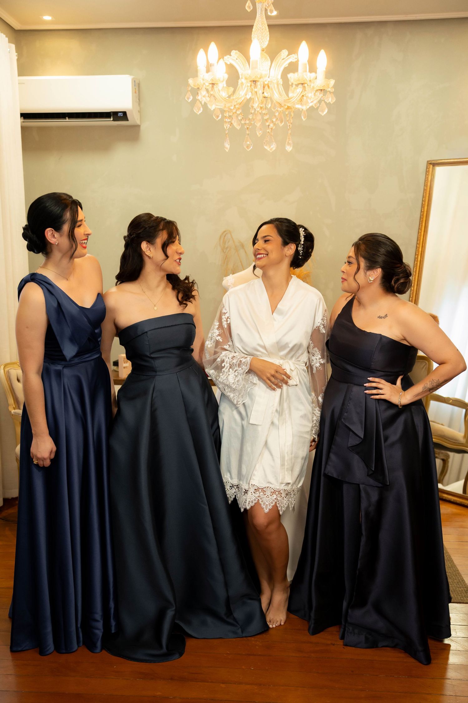 Bride in white robe stands with three bridesmaids in navy blue gowns, smiling together under a chandelier.