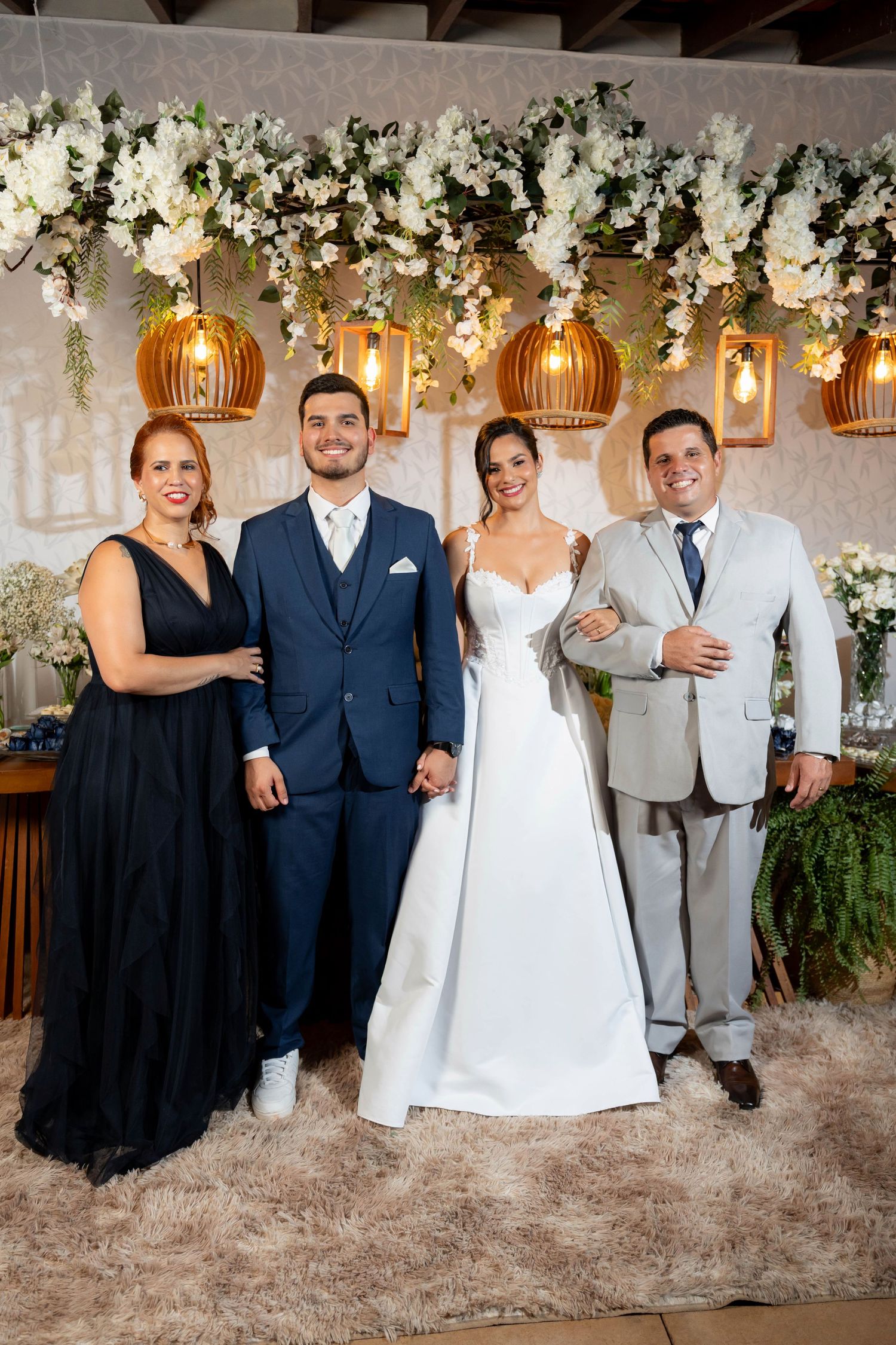 Wedding party of four posing elegantly before floral backdrop with hanging lanterns, bride in white gown, groom in navy suit.