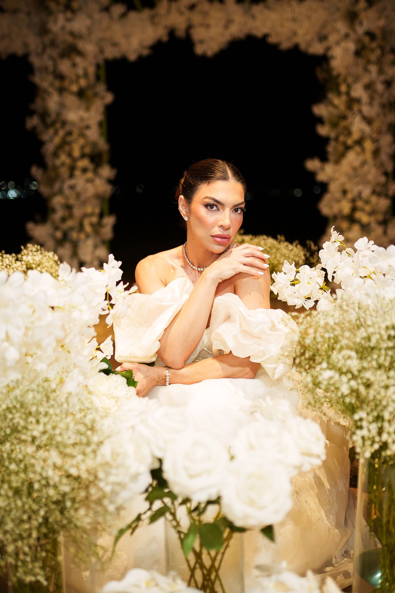 Elegant bride in white ruffled gown surrounded by lush white floral arrangements at a romantic nighttime wedding setting.