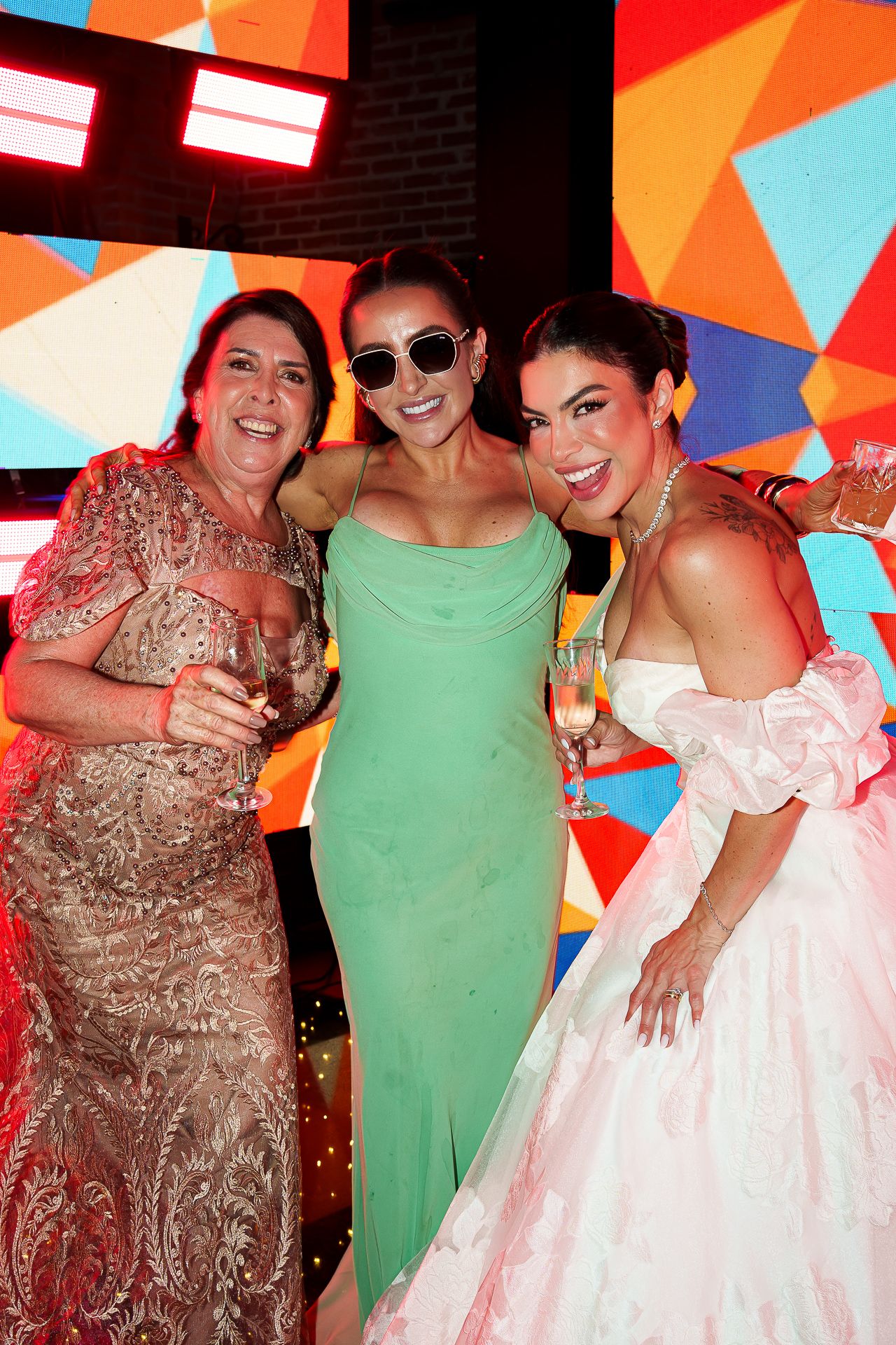 Three women in elegant gowns smile together at a colorful event, holding champagne glasses.