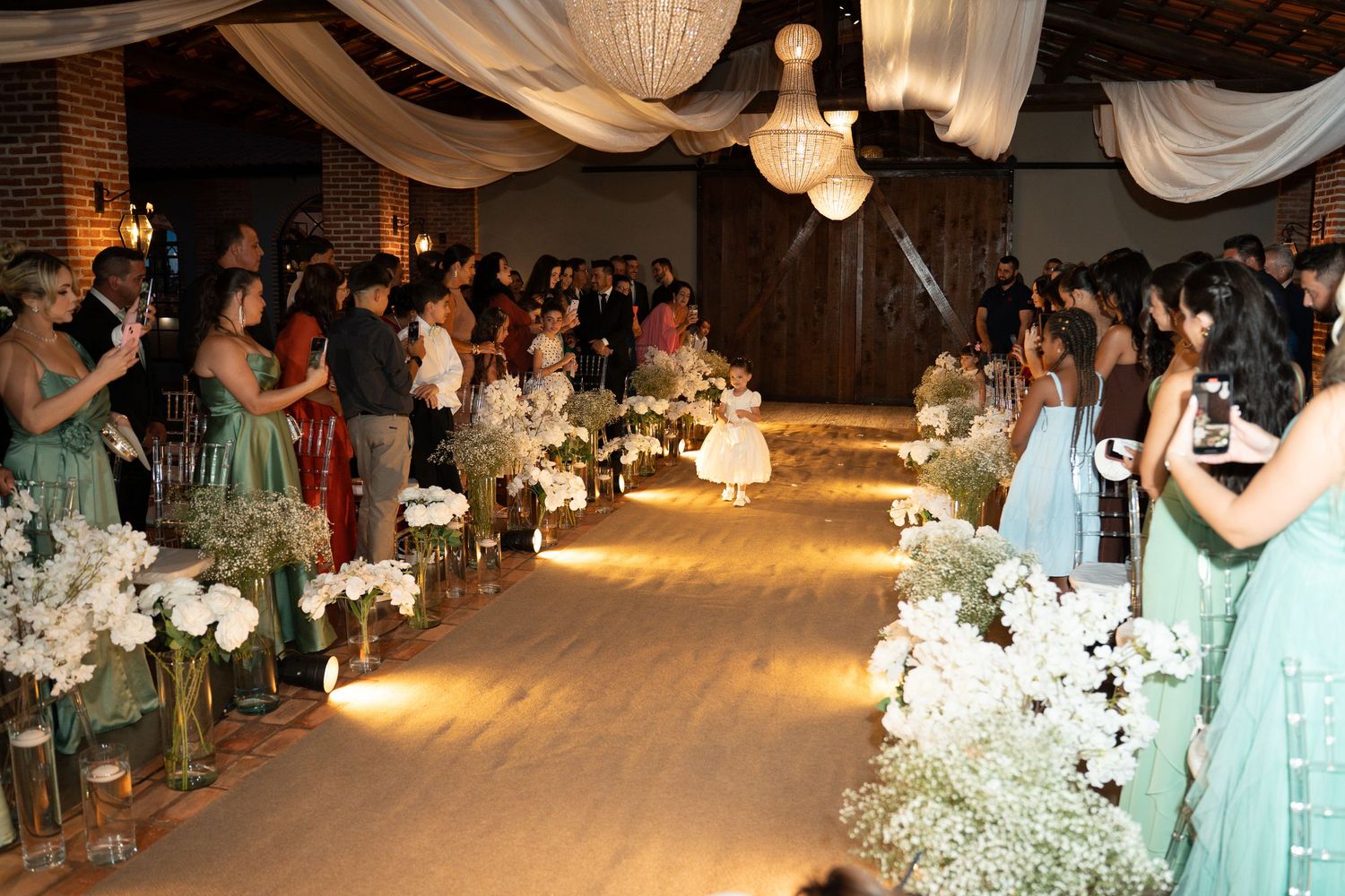 Elegant wedding ceremony aisle lined with white floral arrangements and candles in a decorated indoor venue.