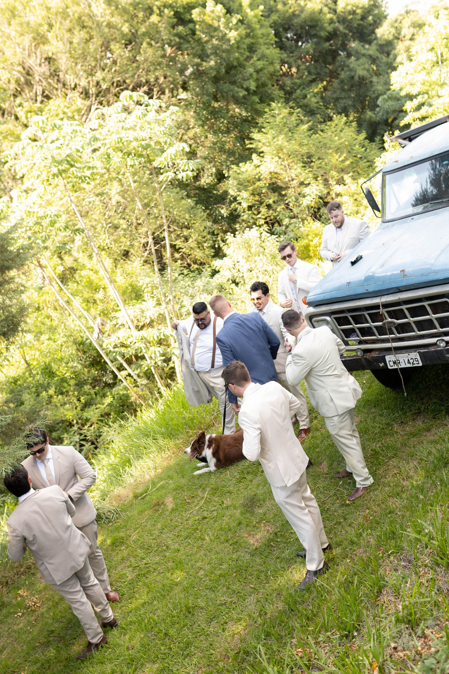 Groomsmen in light suits gather around a vintage blue truck on a grassy hillside with a dog during an outdoor wedding.