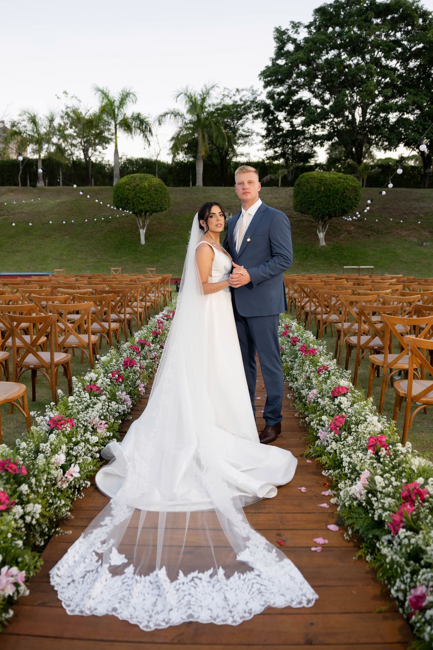 Bride and groom pose together on a floral-lined aisle at an outdoor wedding ceremony with wooden chairs.