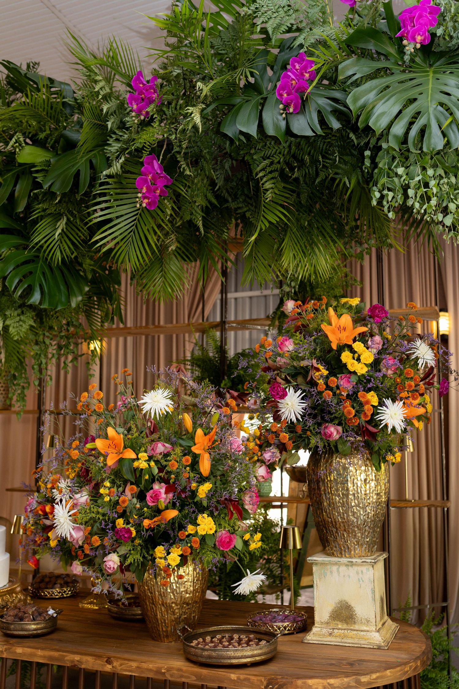 Lush tropical floral arrangements in gold vases with orange lilies, pink blooms, and white flowers under green palm leaves.