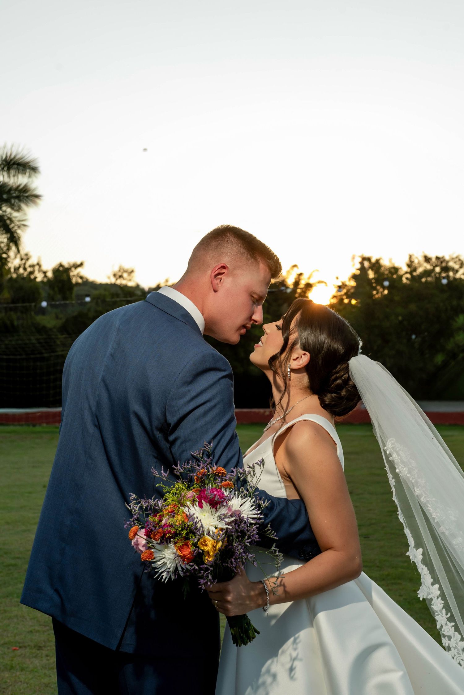 Bride and groom share a romantic moment at sunset, bride holding colorful bouquet wearing veil, groom in blue suit.
