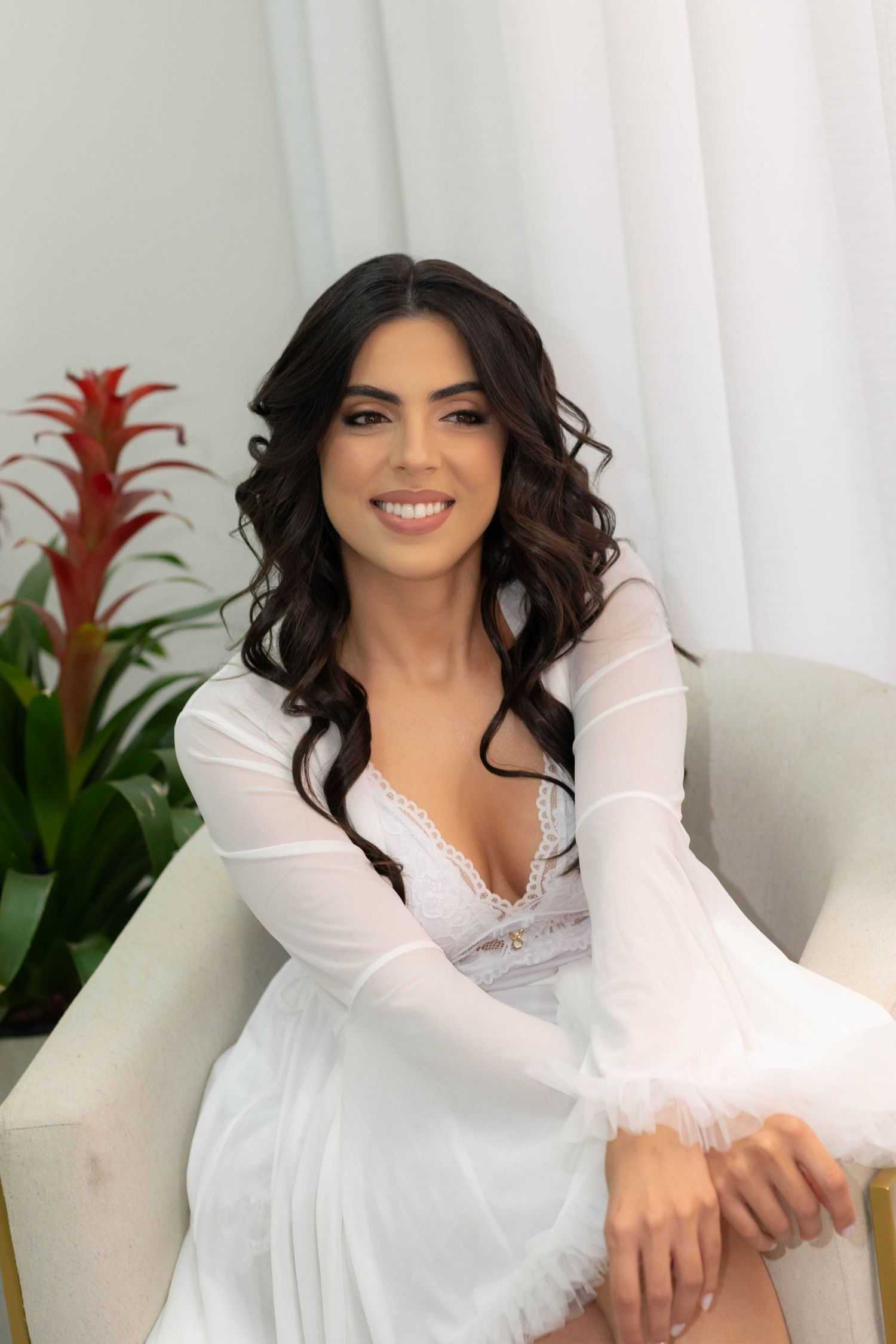 A smiling brunette woman in a white lace dress sits elegantly in a cream chair beside a red tropical plant.