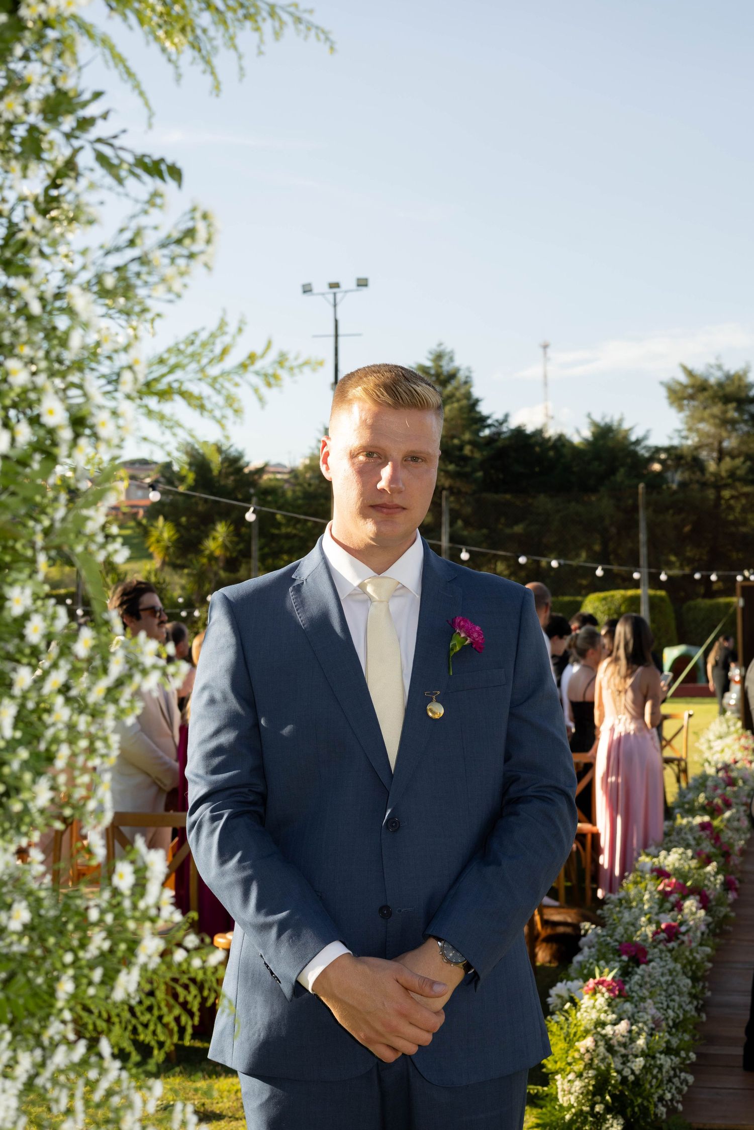 Groom in navy blue suit with pink boutonniere stands at outdoor wedding ceremony surrounded by floral arrangements.