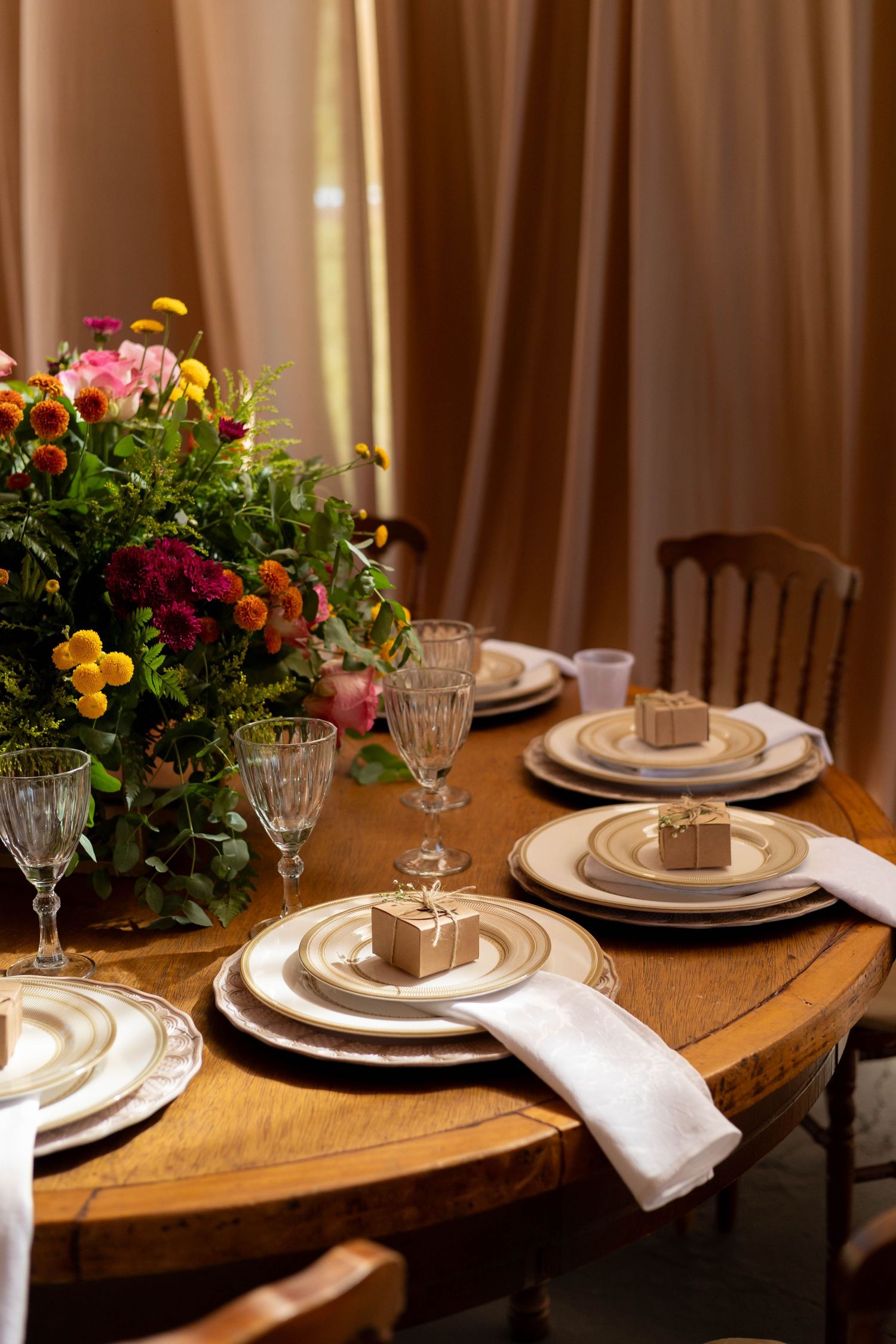 Elegant round wooden dining table set with gold-rimmed plates, crystal glasses, white napkins, and colorful floral centerpiece.