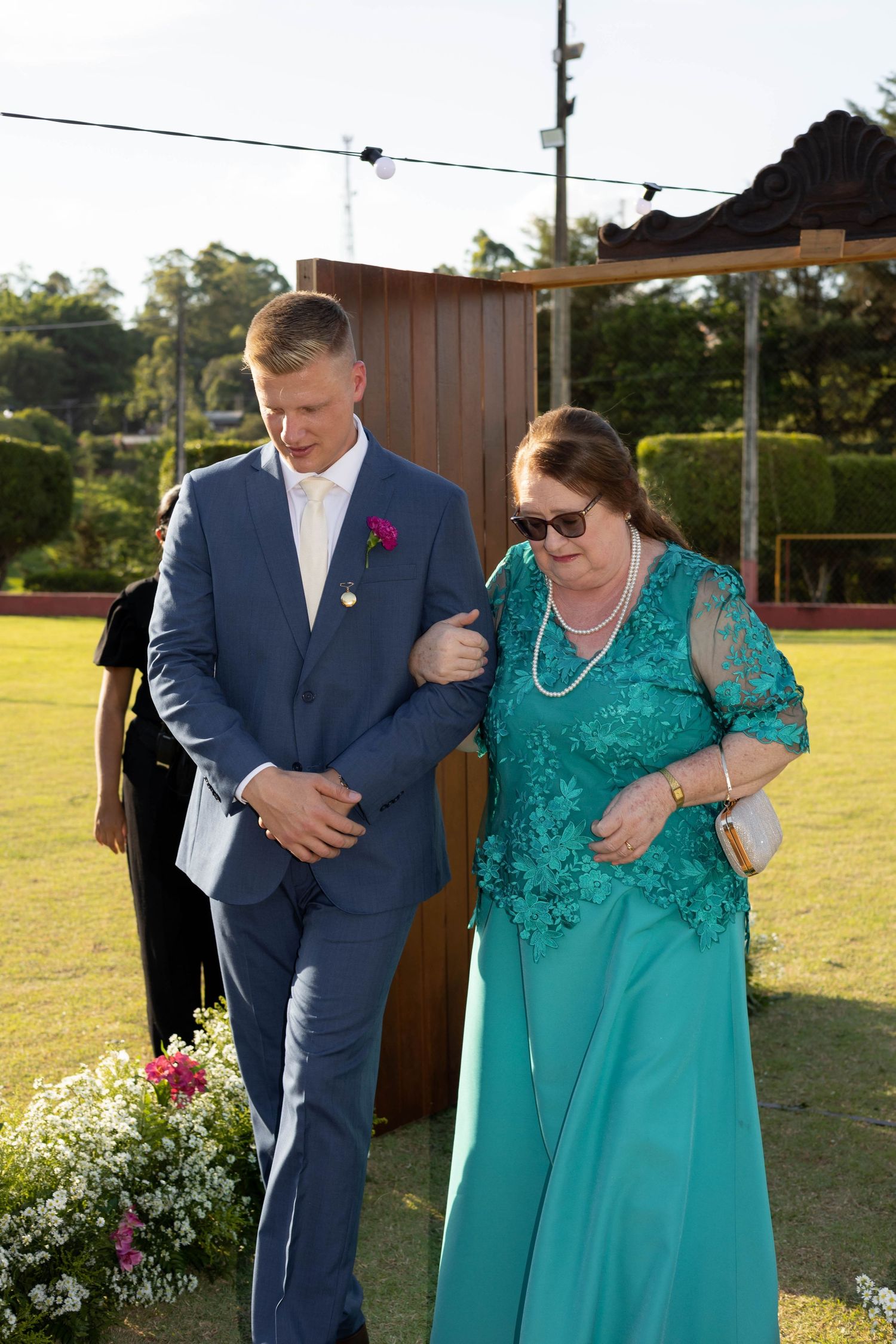 Groom in blue suit escorts elegant woman in teal lace gown at outdoor wedding ceremony on green lawn.