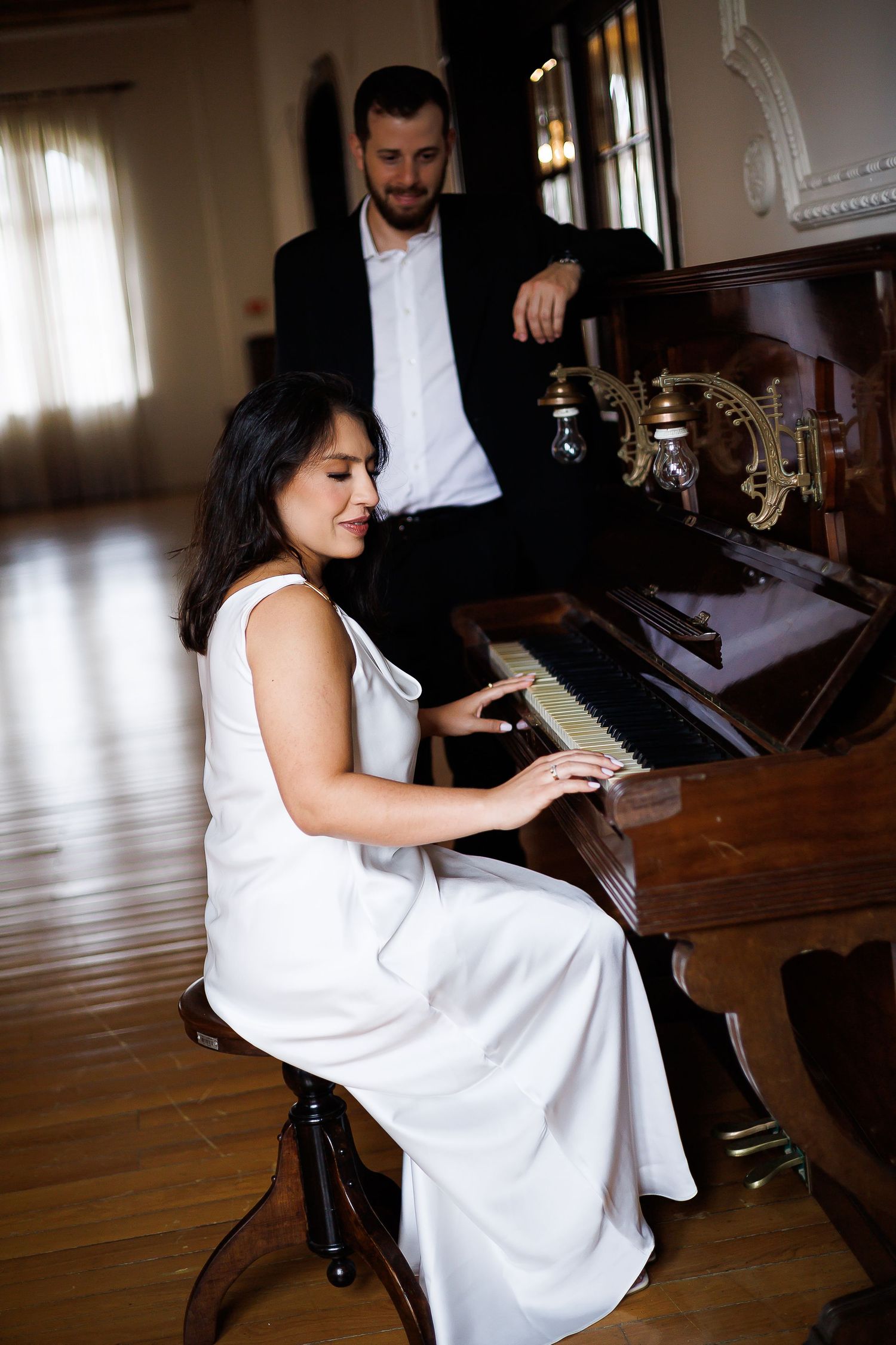 A woman in a white dress plays an upright piano while a man in a black suit stands beside her watching.