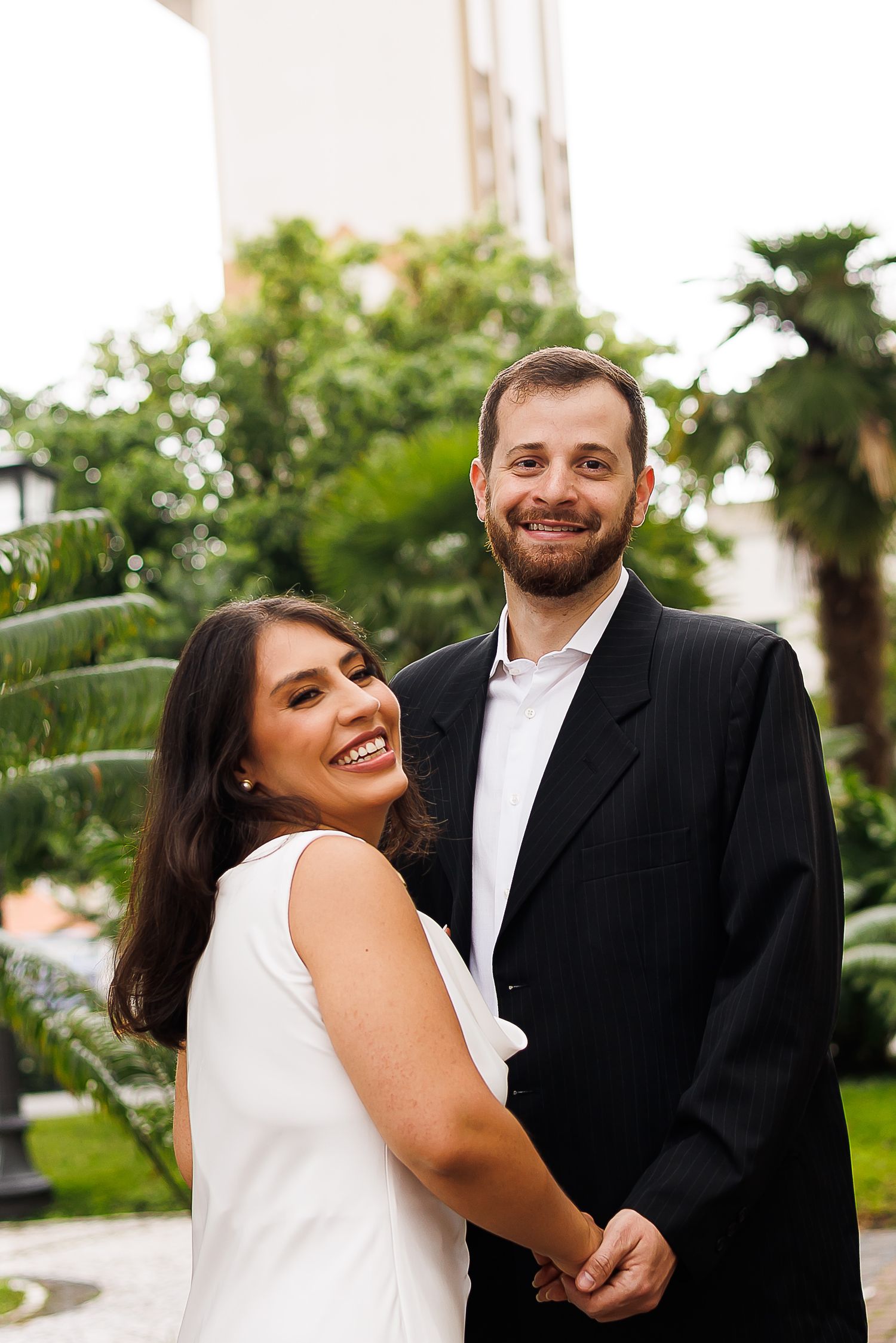 Smiling couple in formal attire posing outdoors with lush green tropical plants and a building in the background.