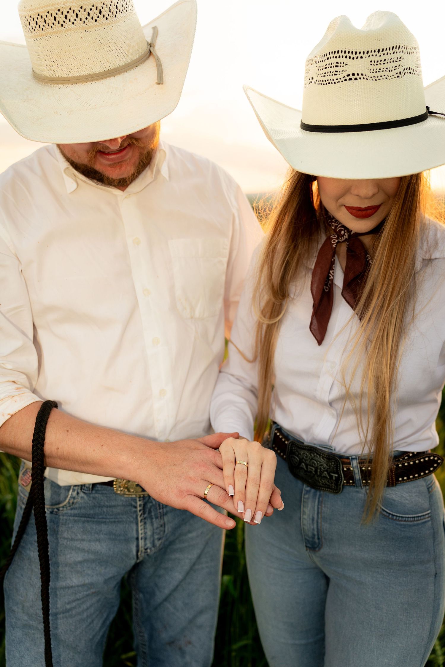 Couple in matching white cowboy hats and jeans holding hands, showing an engagement ring outdoors.