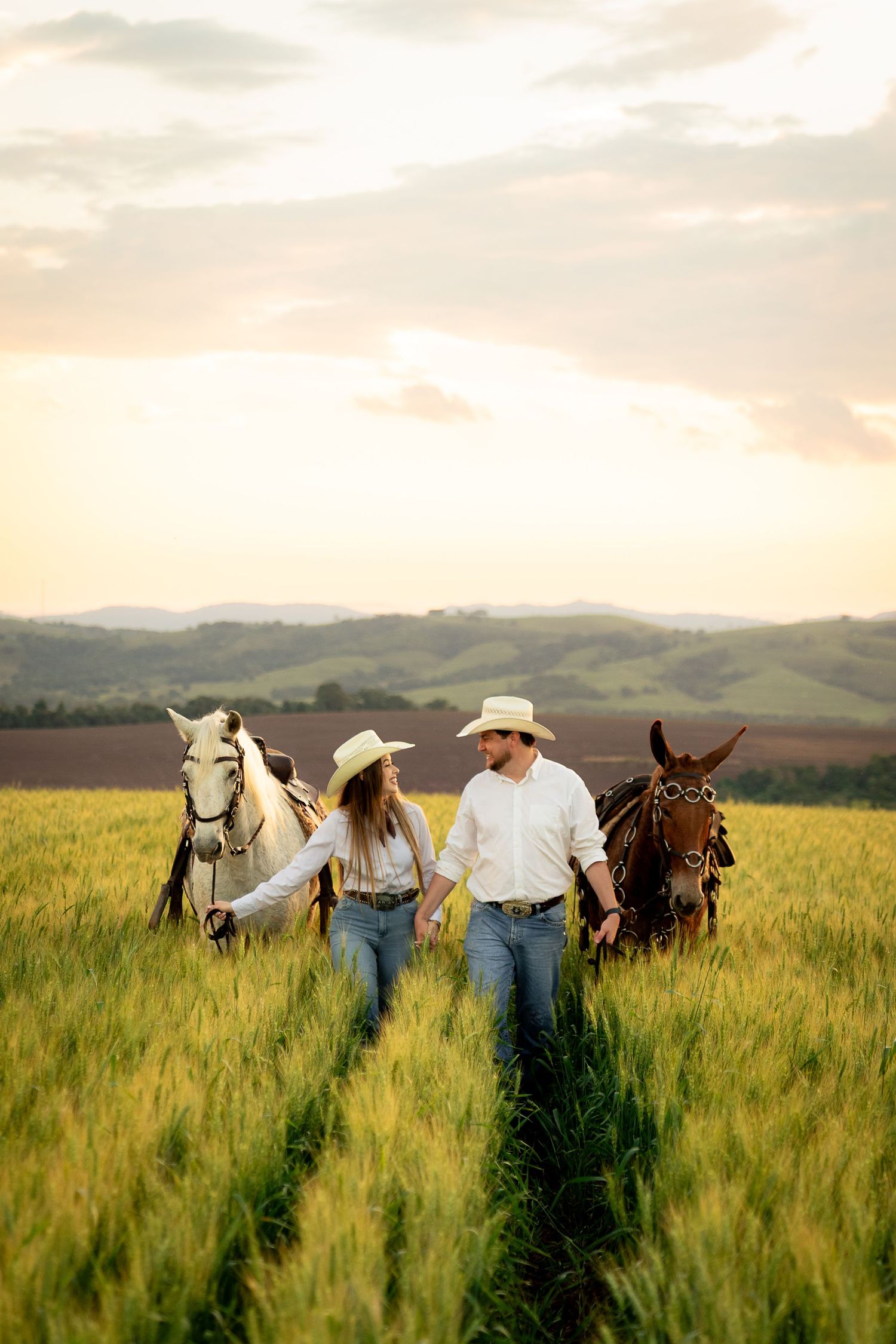 A couple in cowboy hats walks hand-in-hand through a green field at sunset, leading a white and brown horse.
