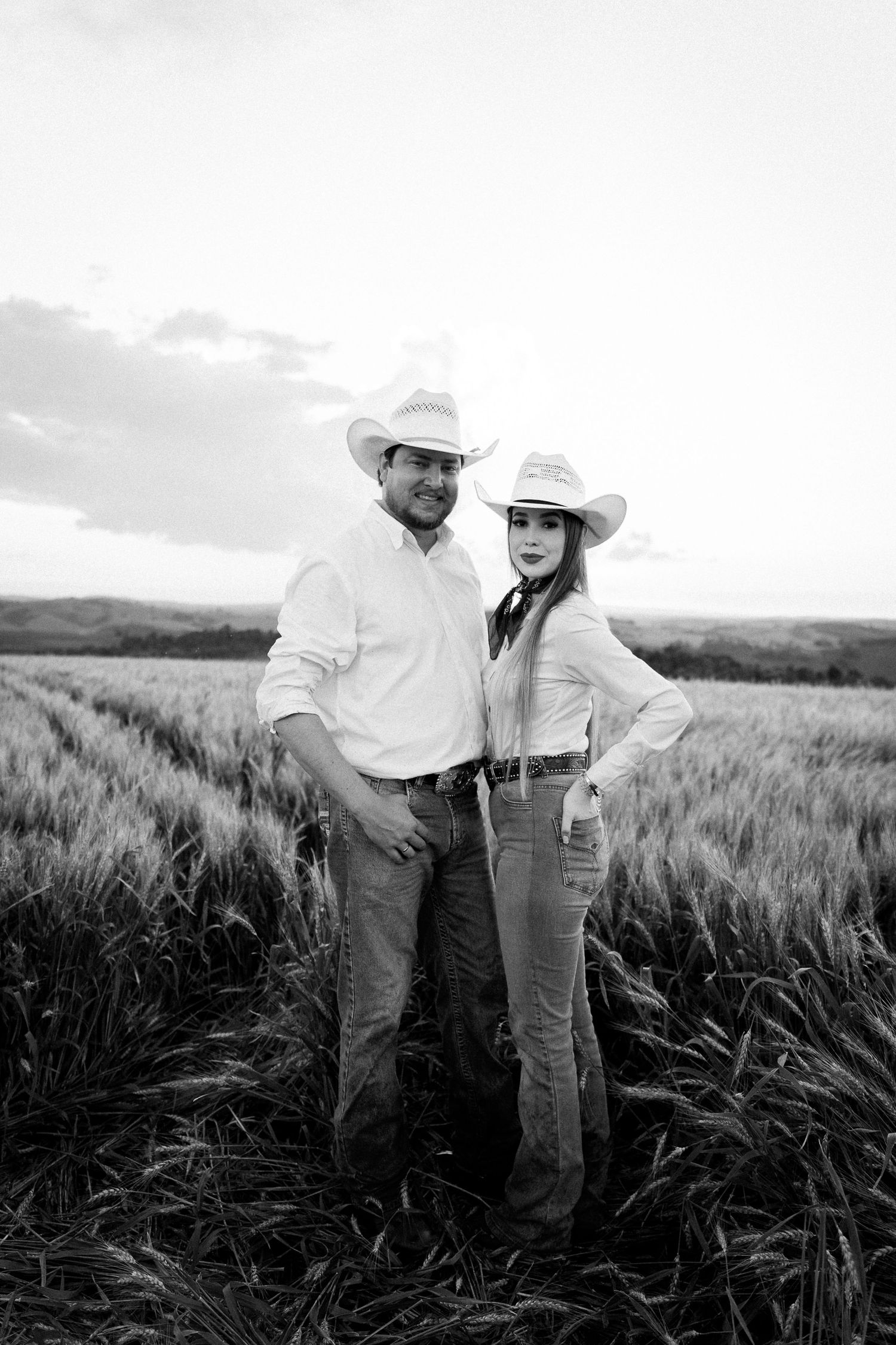 Black and white photo of a couple in cowboy hats posing in a wheat field at sunset.