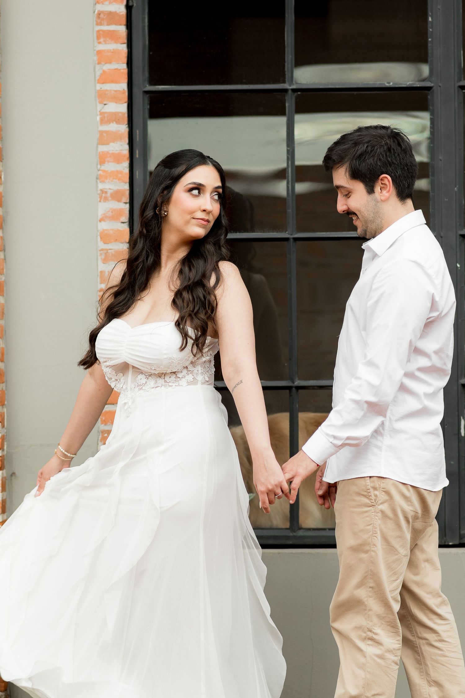 A bride in a flowing white gown holds hands with a groom in a white shirt outdoors near a brick wall.