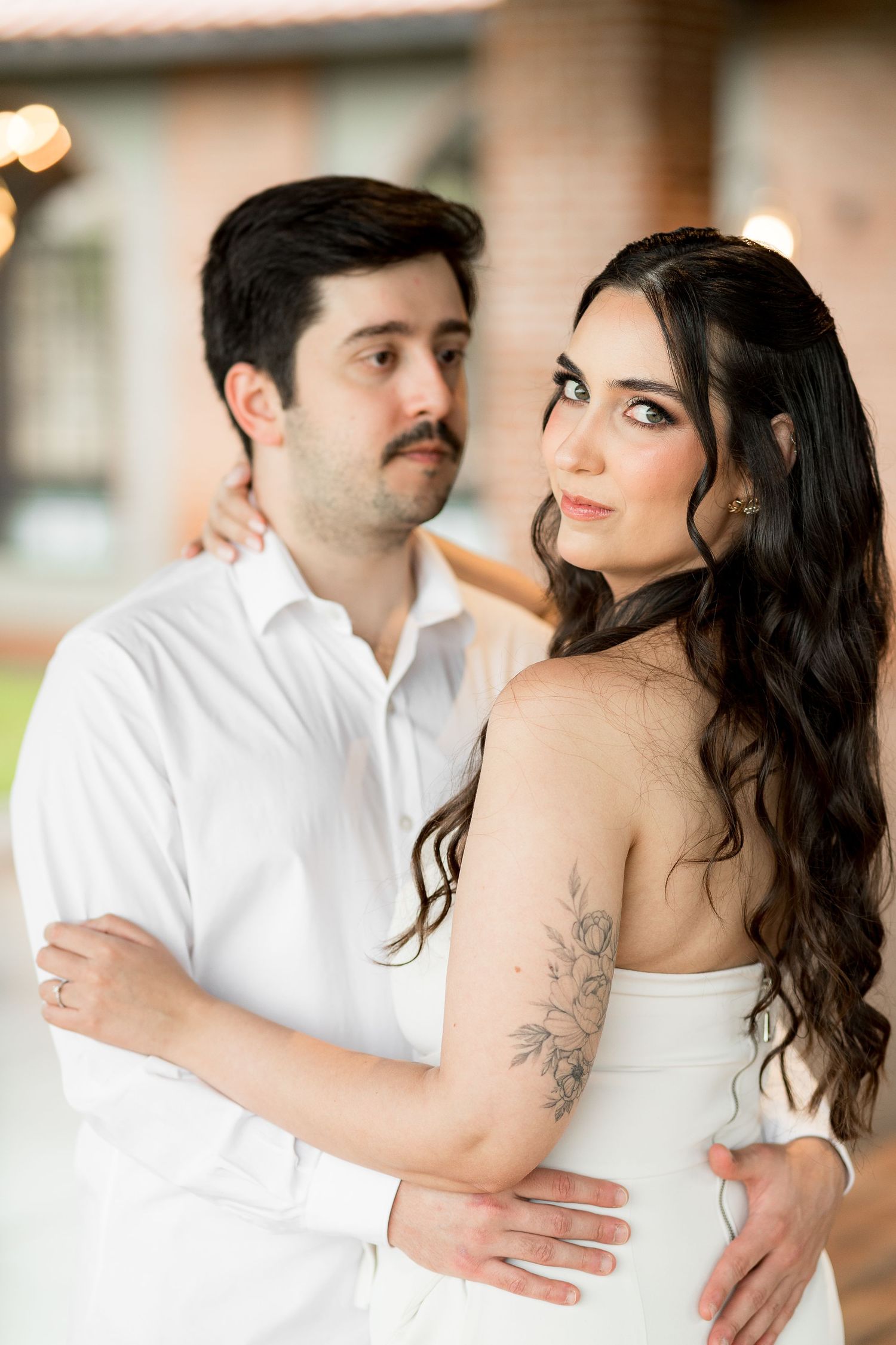 Couple in white attire embracing, woman with floral arm tattoo and wavy dark hair gazes away, man holds her waist.