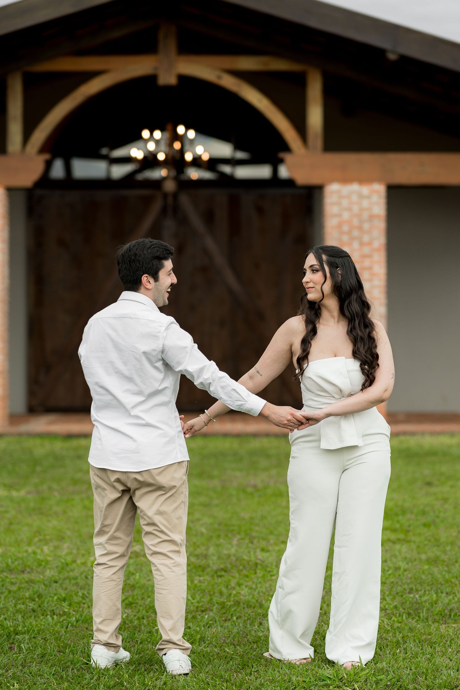 Couple in white outfits holding hands on green lawn before rustic barn with chandelier.