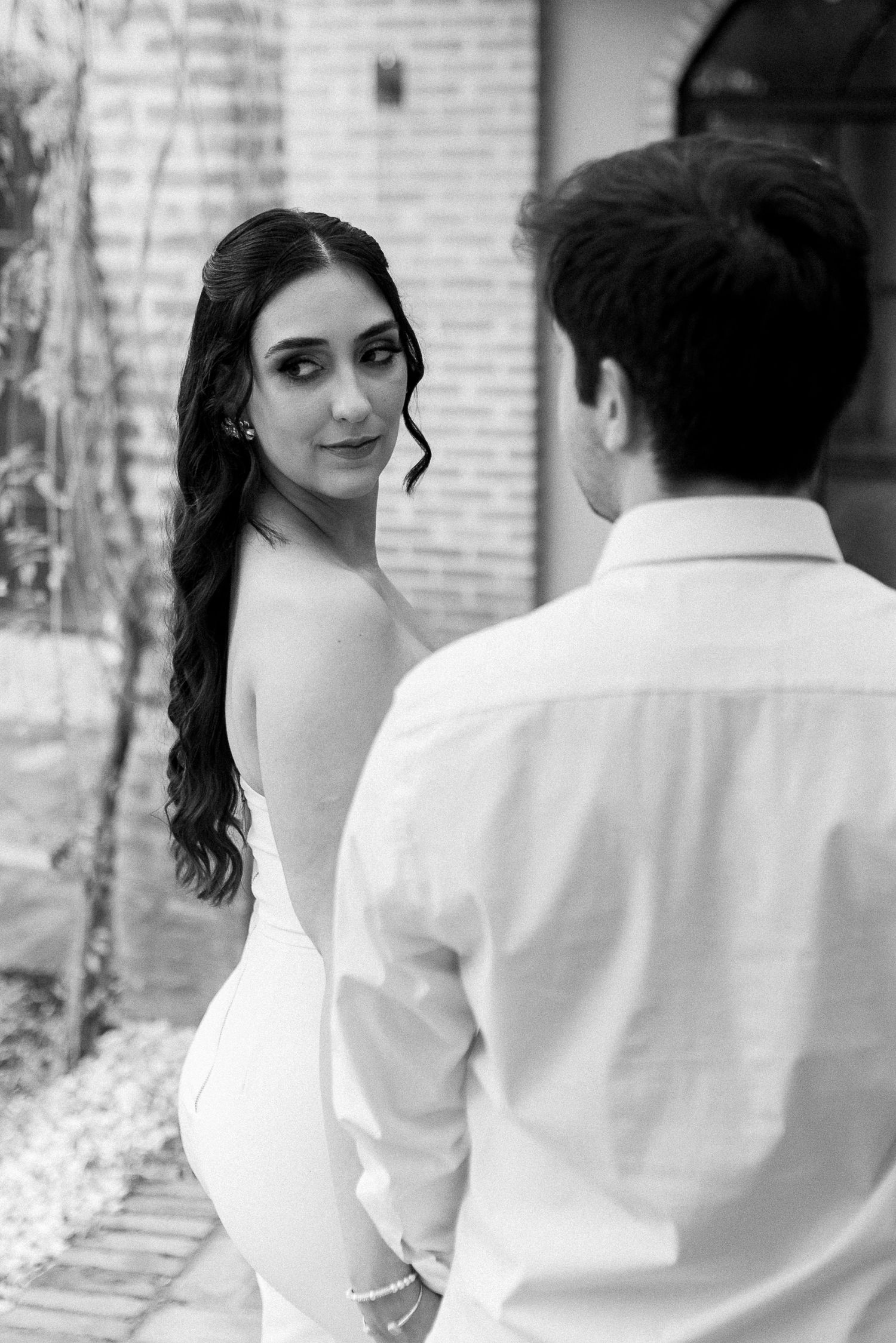 Black and white photo of a bride in a backless gown glancing over her shoulder at a groom in a white shirt.