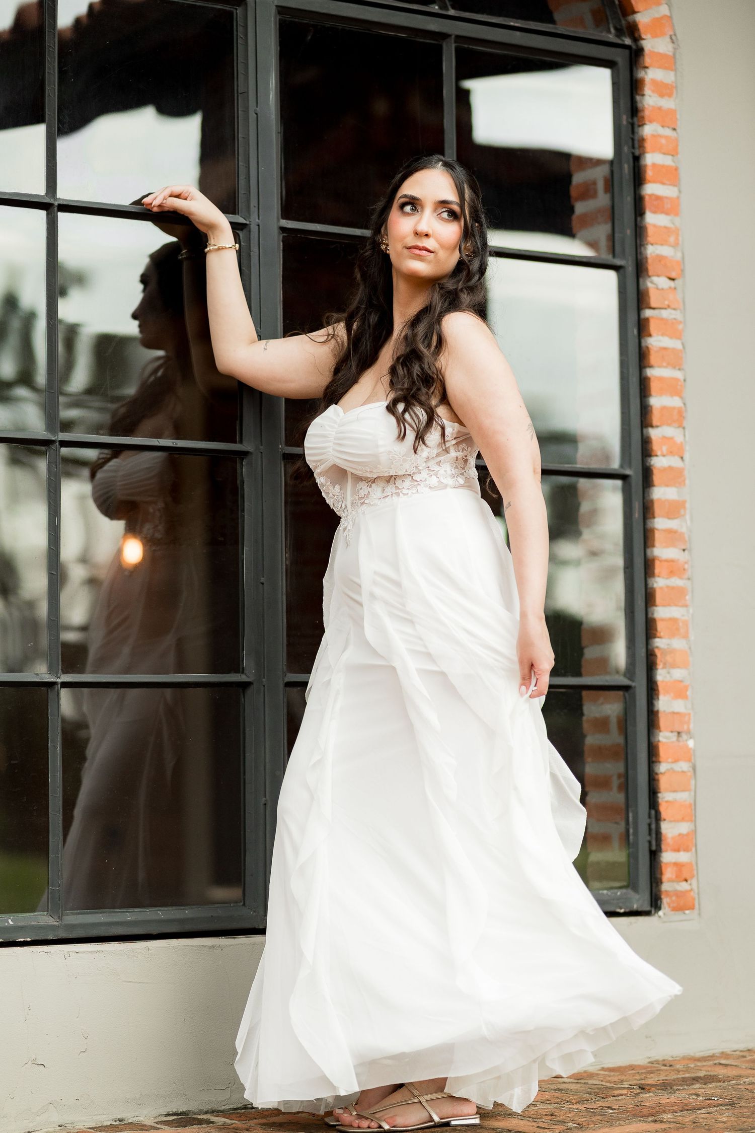Bride in a strapless white flowing wedding gown poses gracefully by large black-framed windows with brick accents.