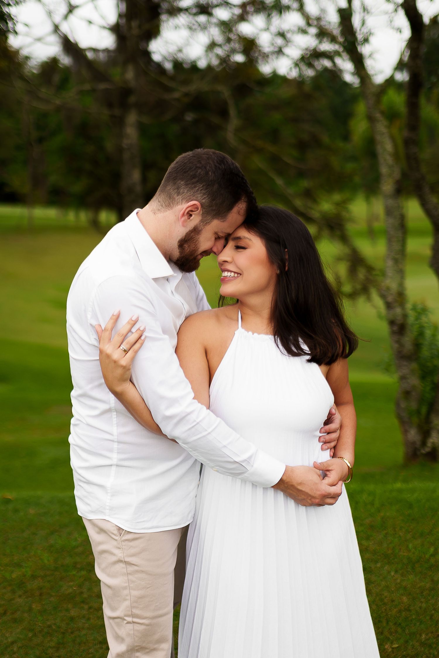 Couple in white outfits embracing outdoors on green lawn, woman smiling joyfully as man leans in close.