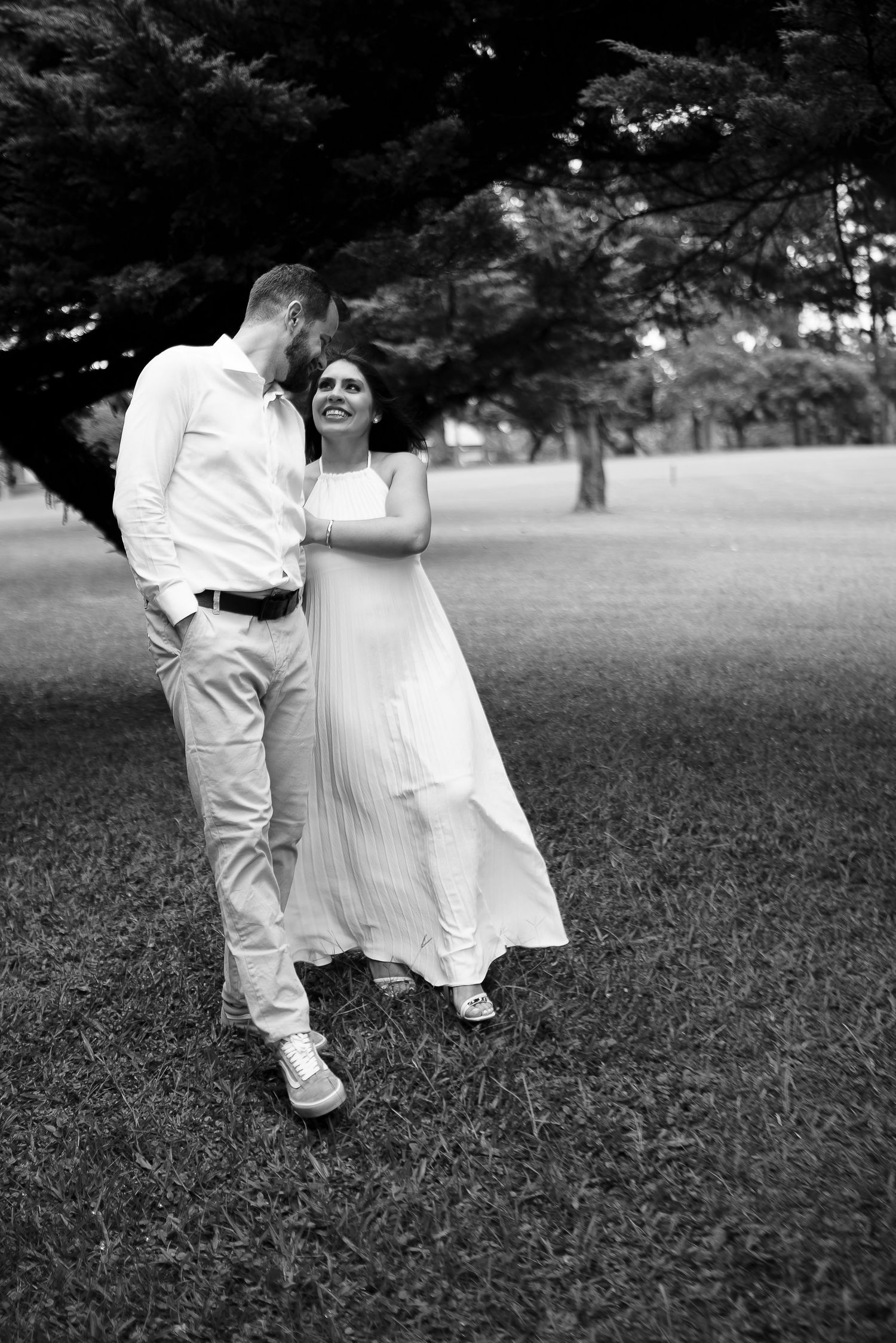 Black and white photo of a couple walking together in a park, woman in a flowing white dress smiling at her partner.