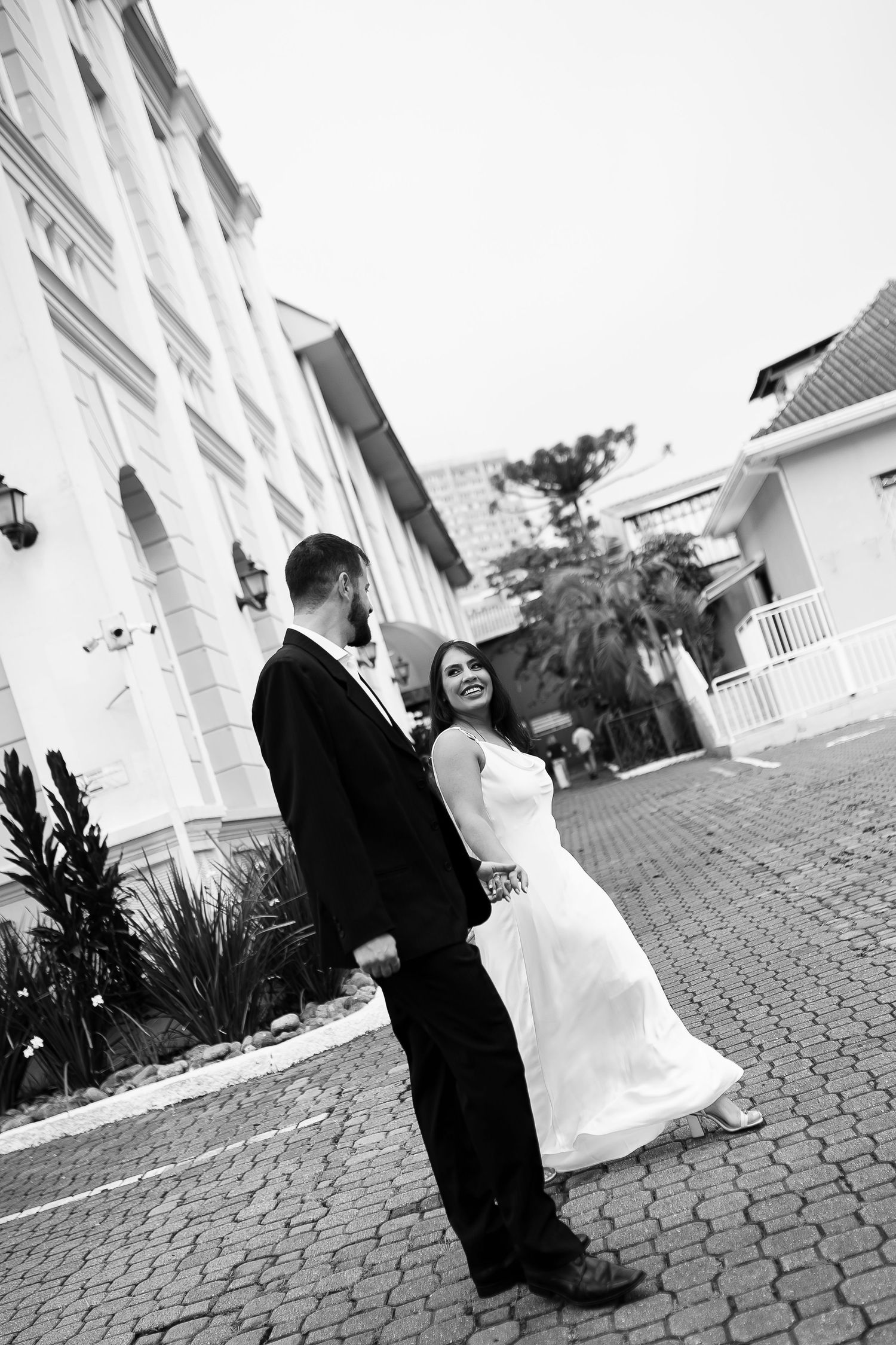 Black and white photo of smiling bride in white gown holding hands with groom in black suit on cobblestone courtyard.