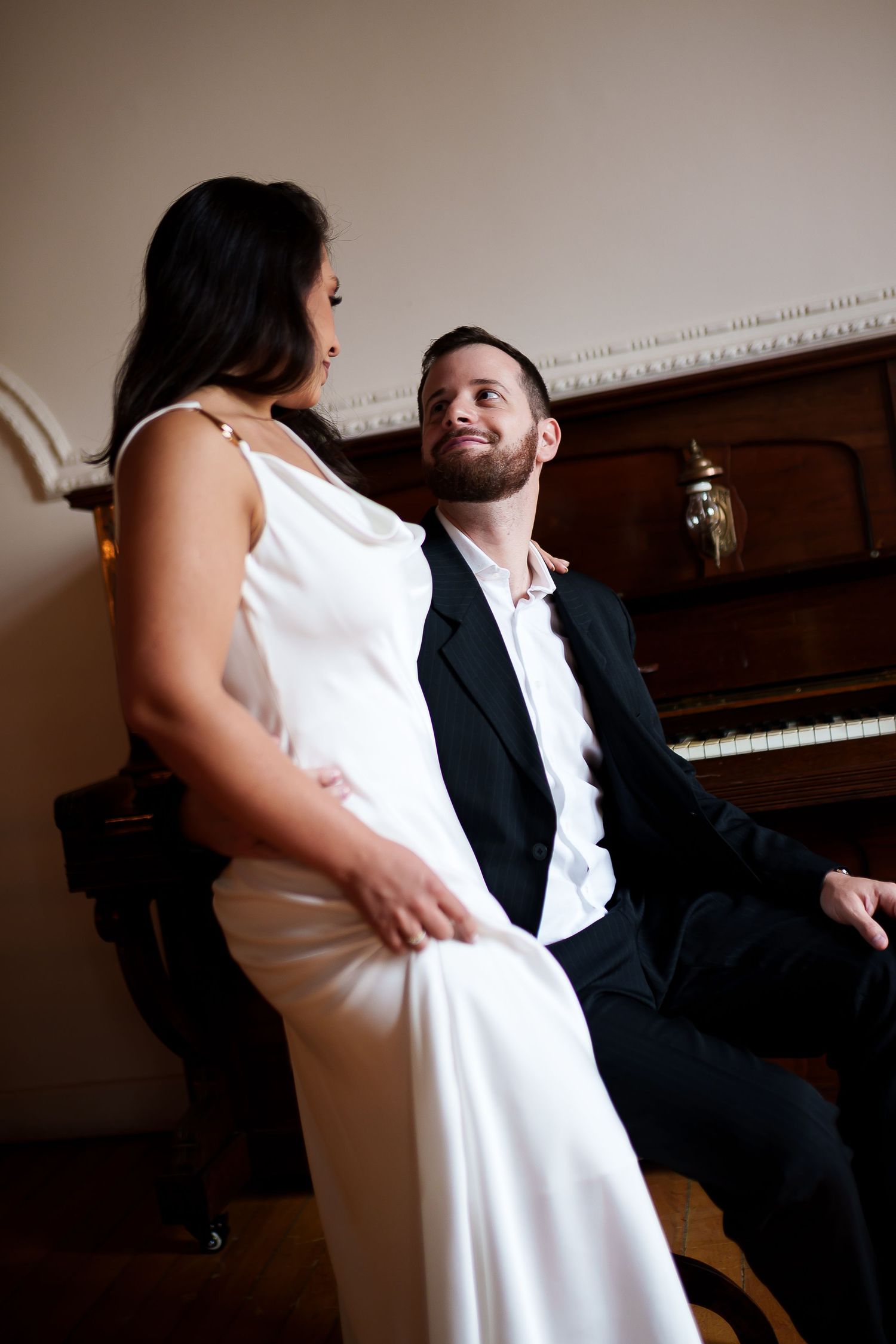 Elegant couple in formal attire sharing a romantic moment by a vintage piano, woman in white gown, man in black suit.