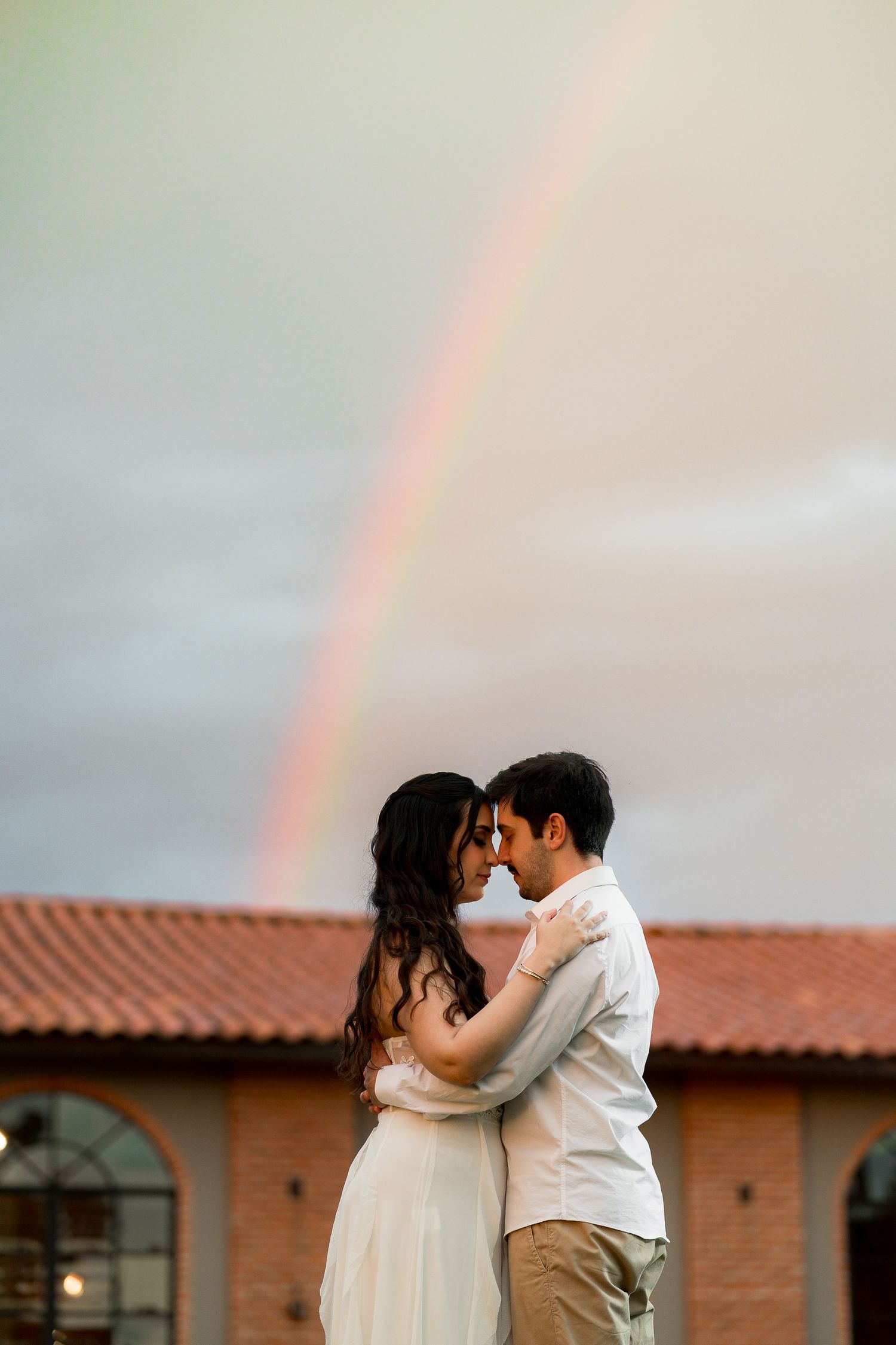 Romantic couple embracing under a beautiful rainbow in a cloudy sky, with a red brick building behind them.