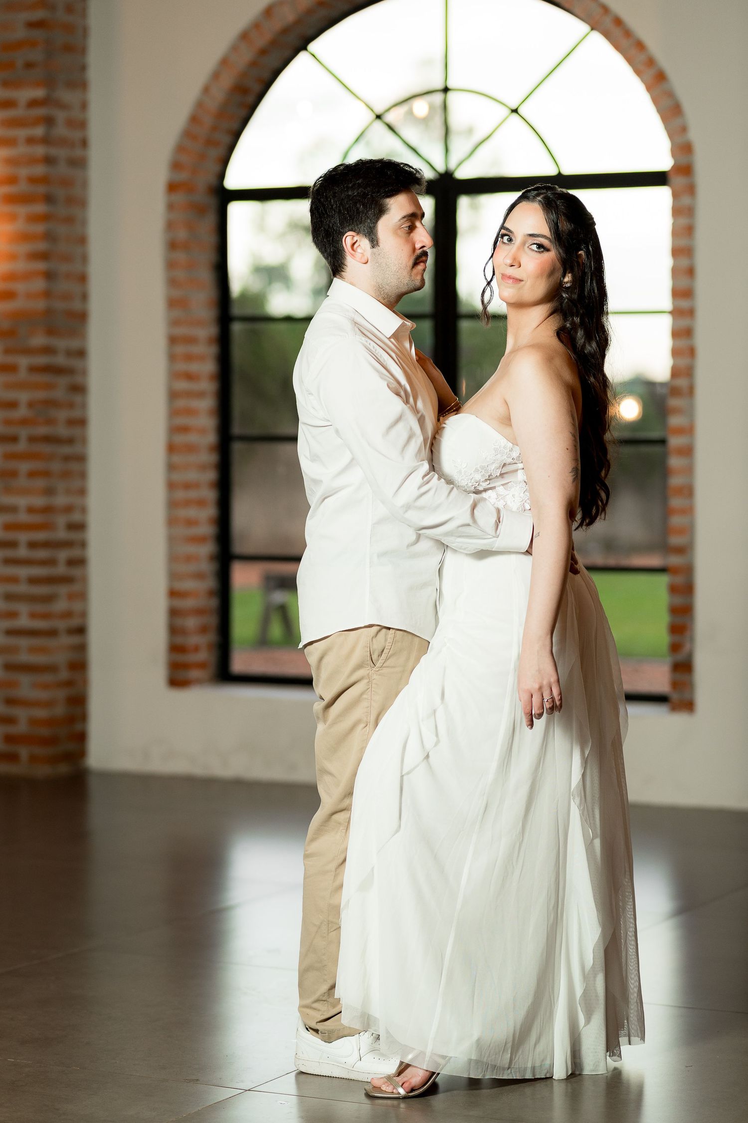 Couple in wedding attire embracing near arched brick window, bride in white gown, groom in tan suit.