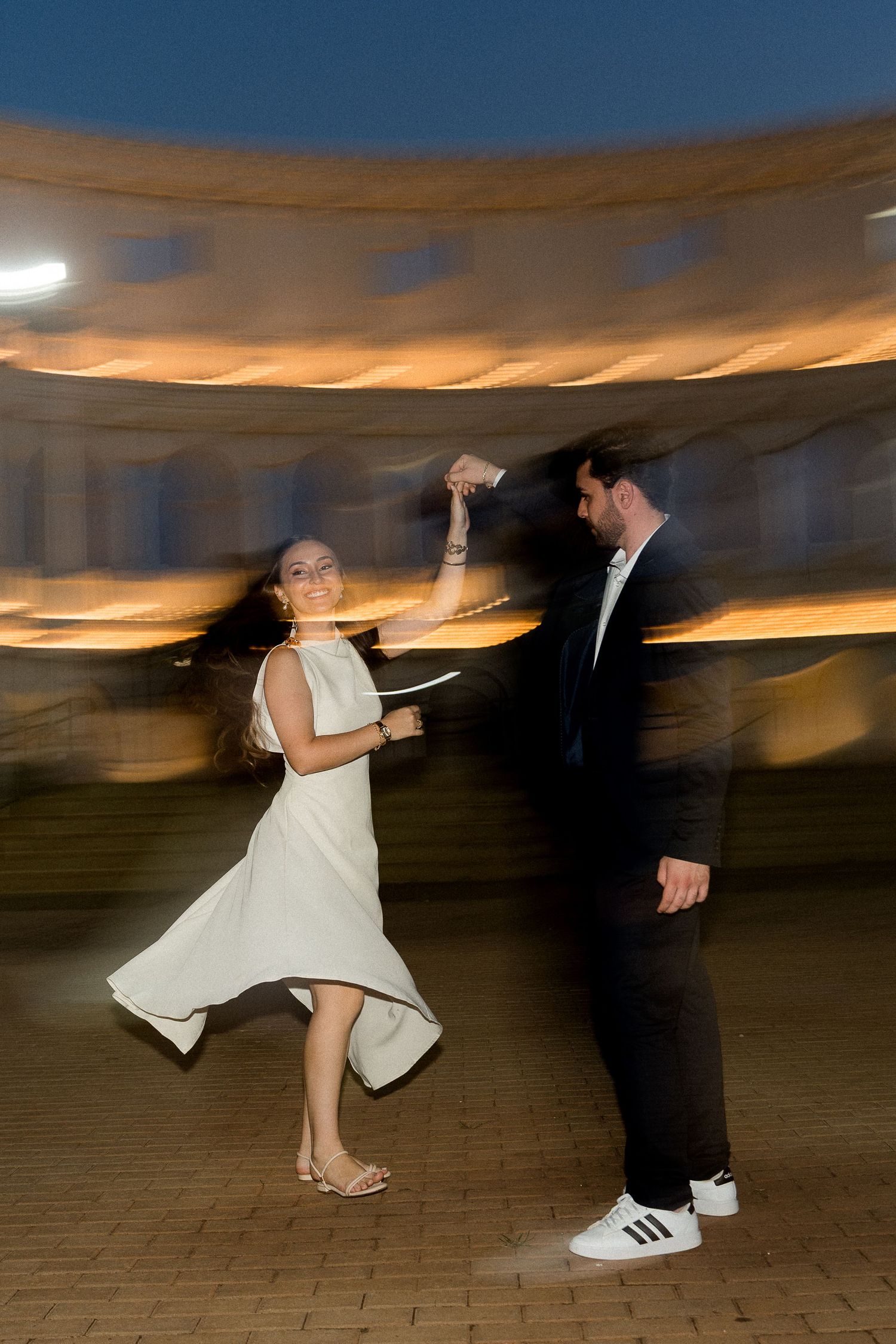 Couple dancing joyfully at dusk, woman in white dress twirling, motion blur capturing energy at an outdoor venue.