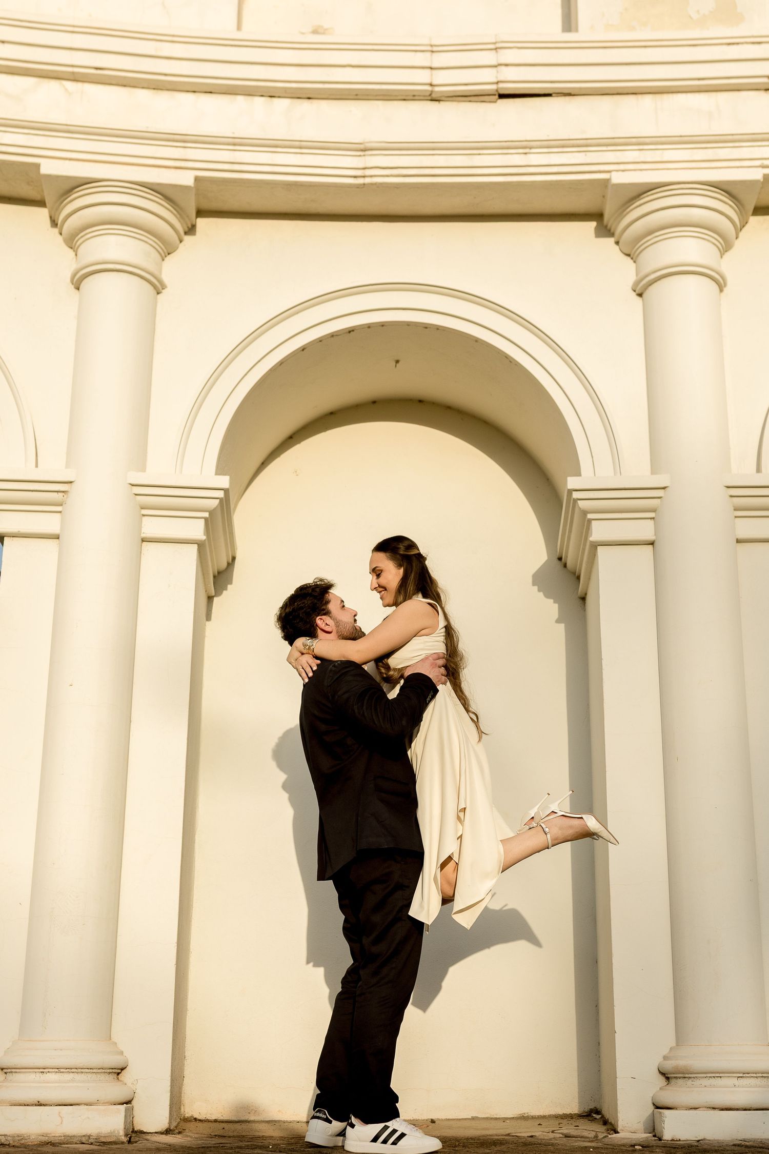 A man in a black suit lifts a woman in a cream dress, both smiling, posed before an elegant white arched building.