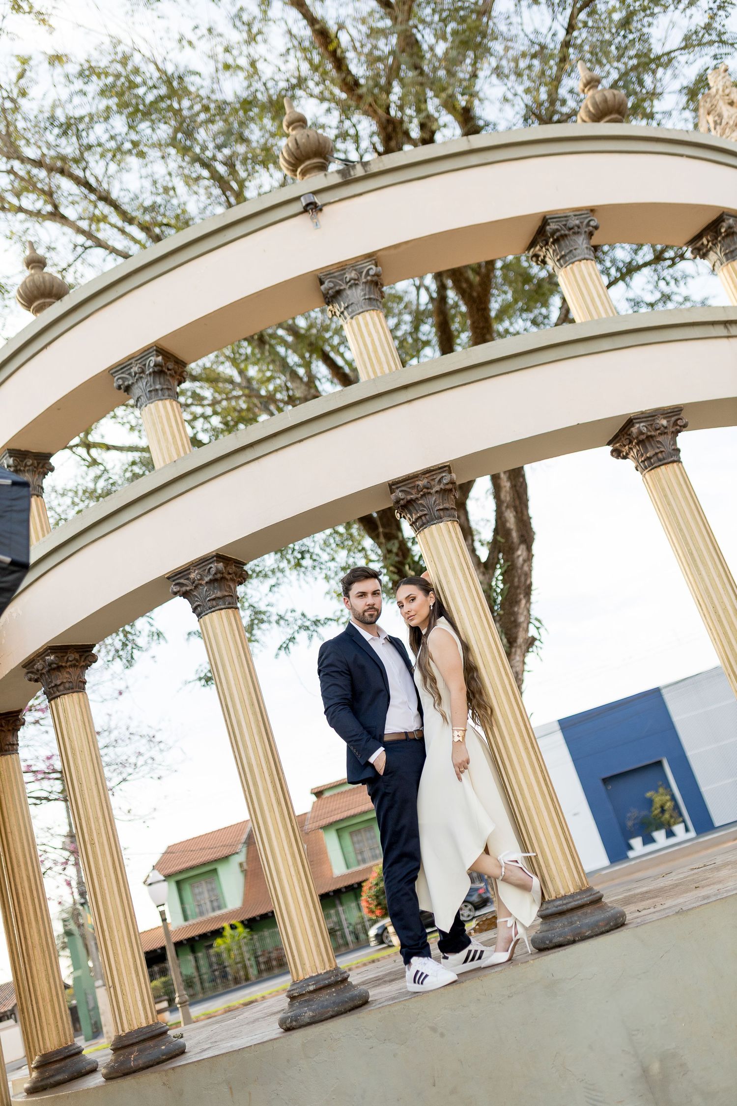 Elegant couple posing under a classical arched pavilion with columns in a park setting.