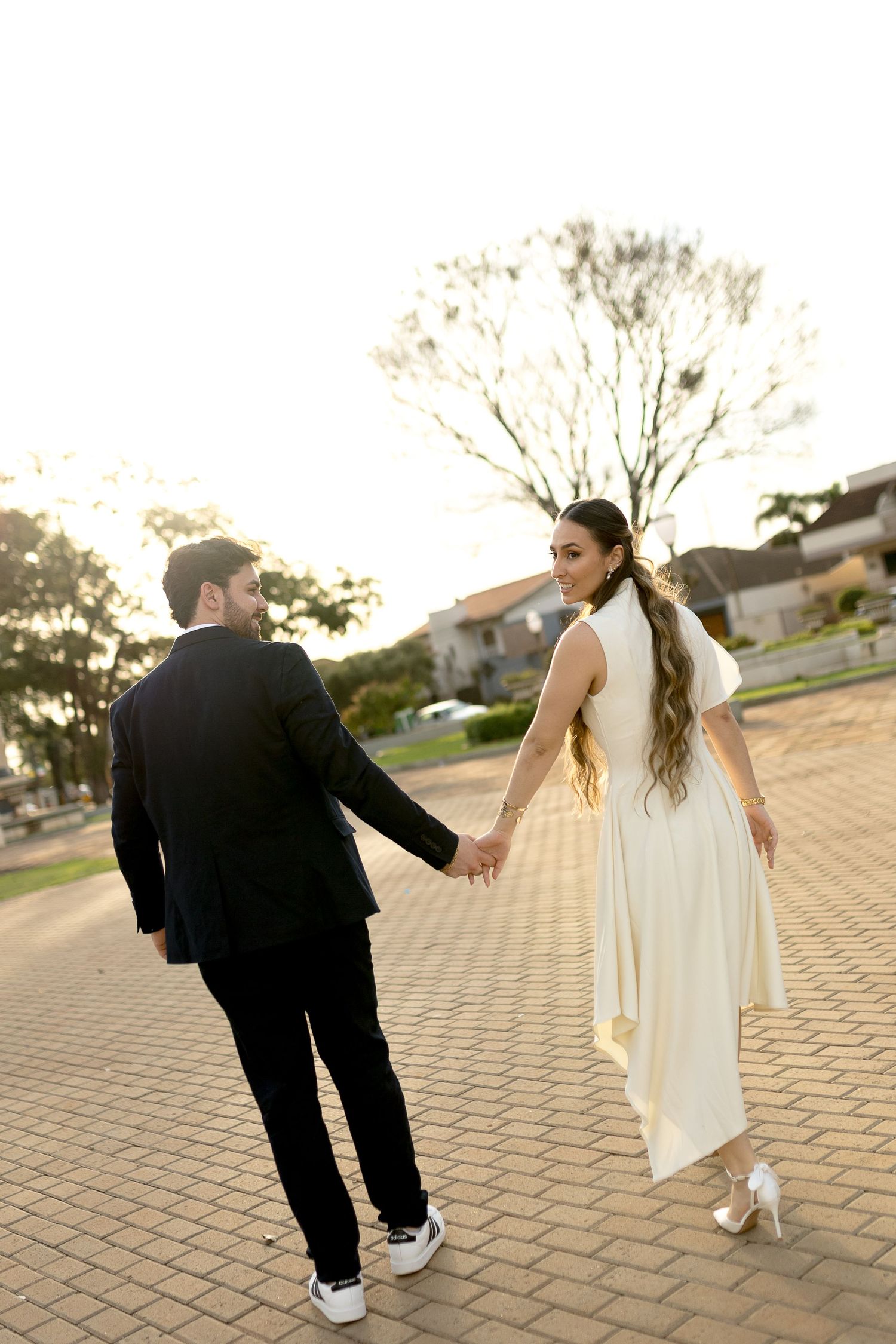 Couple holding hands walking on brick path, woman in white dress looking back, man in black suit, outdoor golden hour setting.