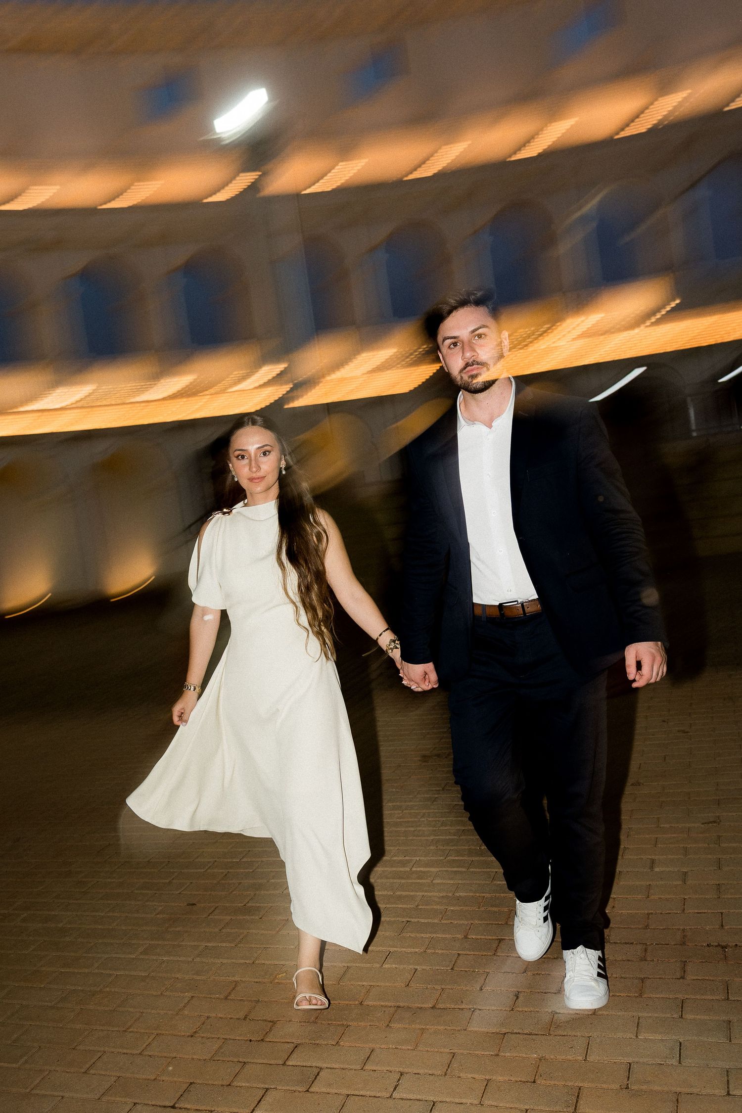 Couple holding hands walking together at night, woman in white dress and man in black suit under warm lights.