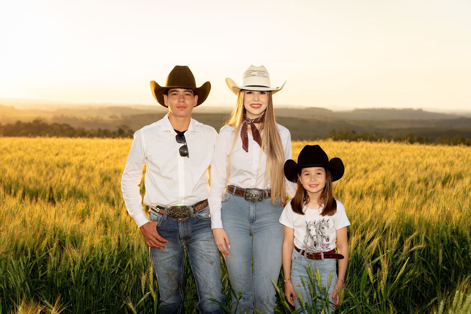 Three people in cowboy hats posing in a golden wheat field at sunset.