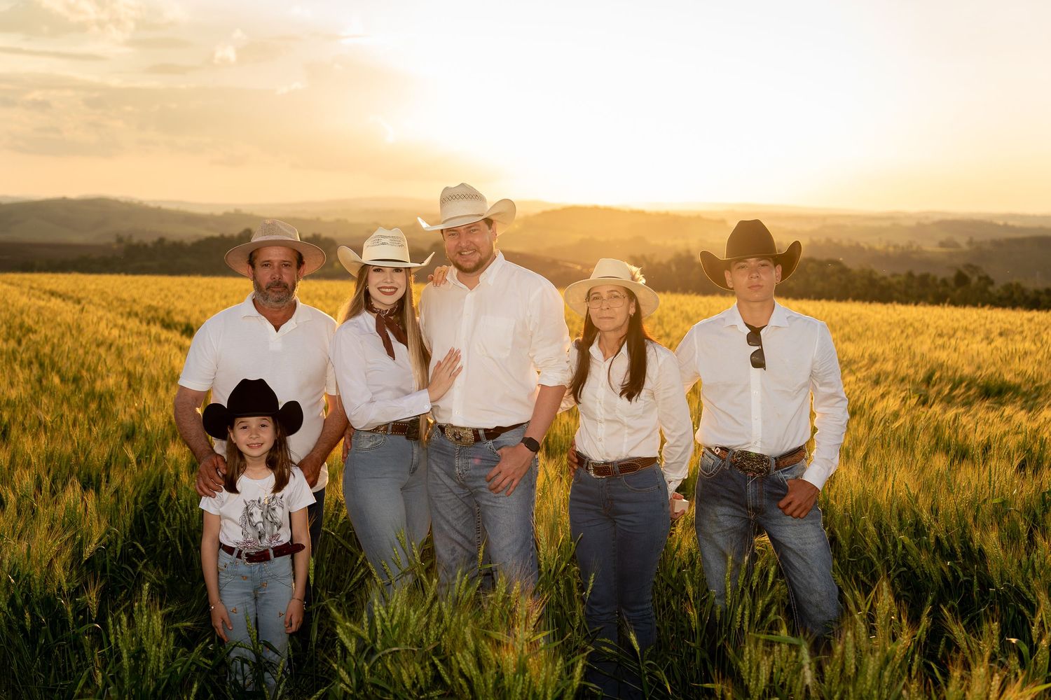 Family in white shirts, jeans, and cowboy hats posing in a golden wheat field at sunset.
