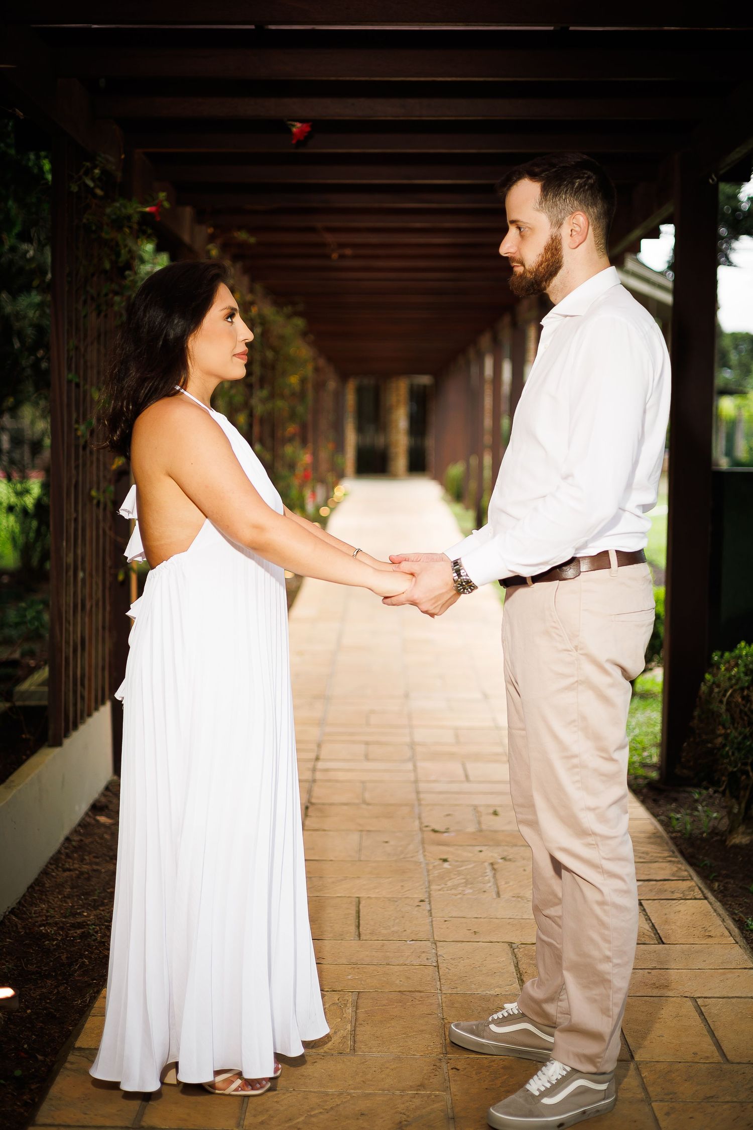 Couple in white dress and casual attire holding hands under wooden pergola on stone pathway.