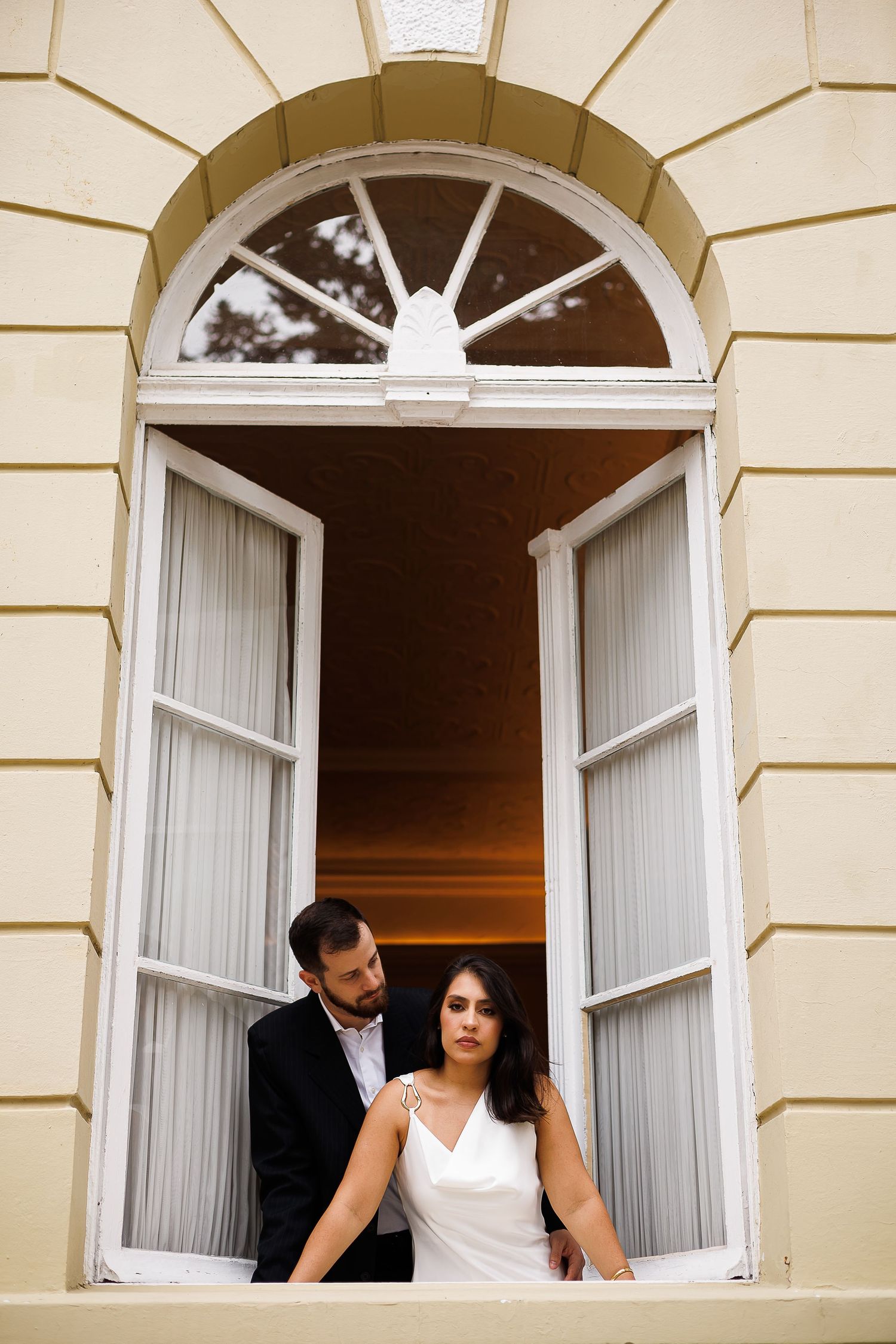 Bride and groom posing romantically in an arched window of an elegant building during their wedding photo shoot.
