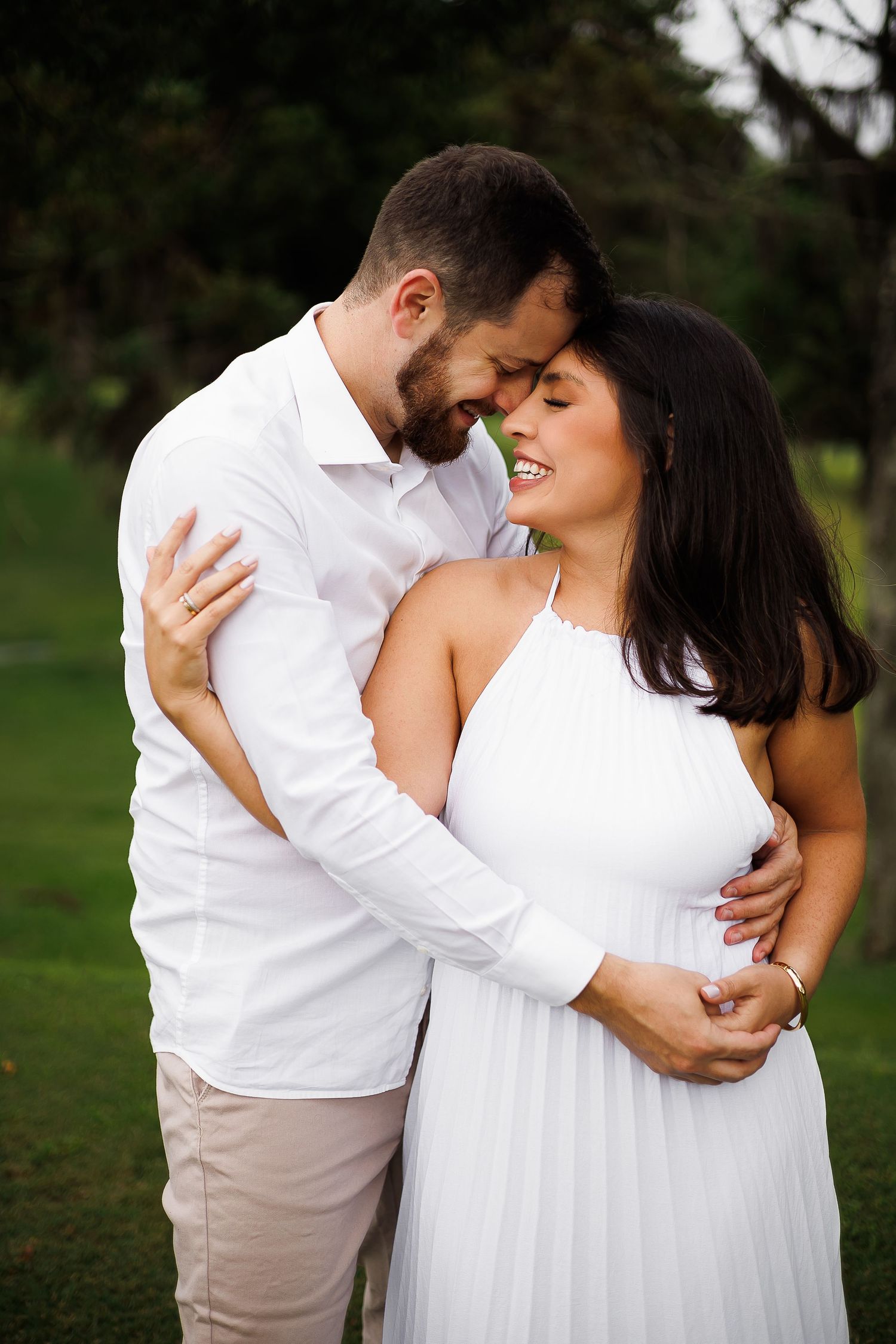 Couple in white outfits embracing outdoors, smiling intimately against a lush green park background.