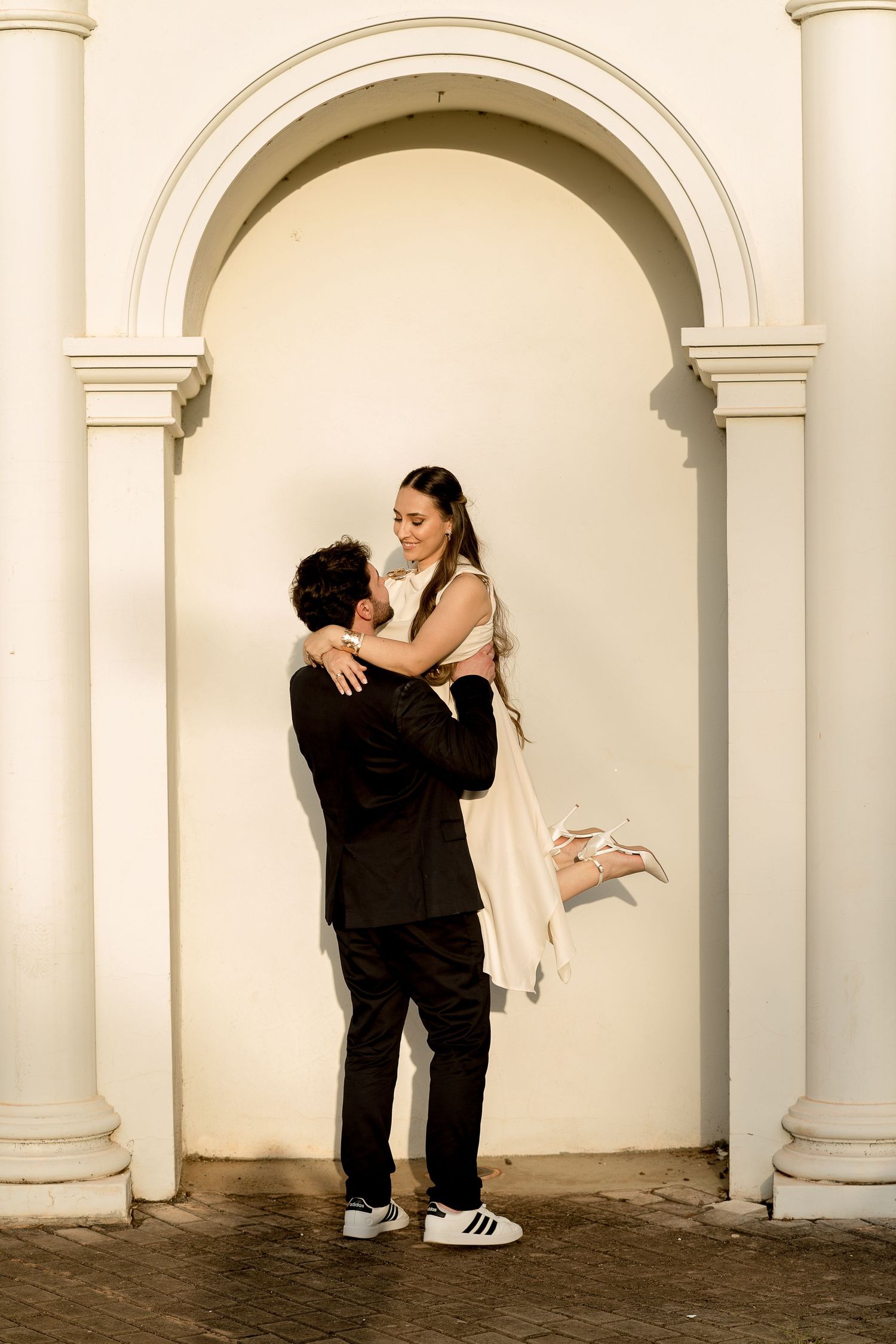 Groom lifting bride in white dress under elegant arch, romantic wedding portrait with white columns backdrop.