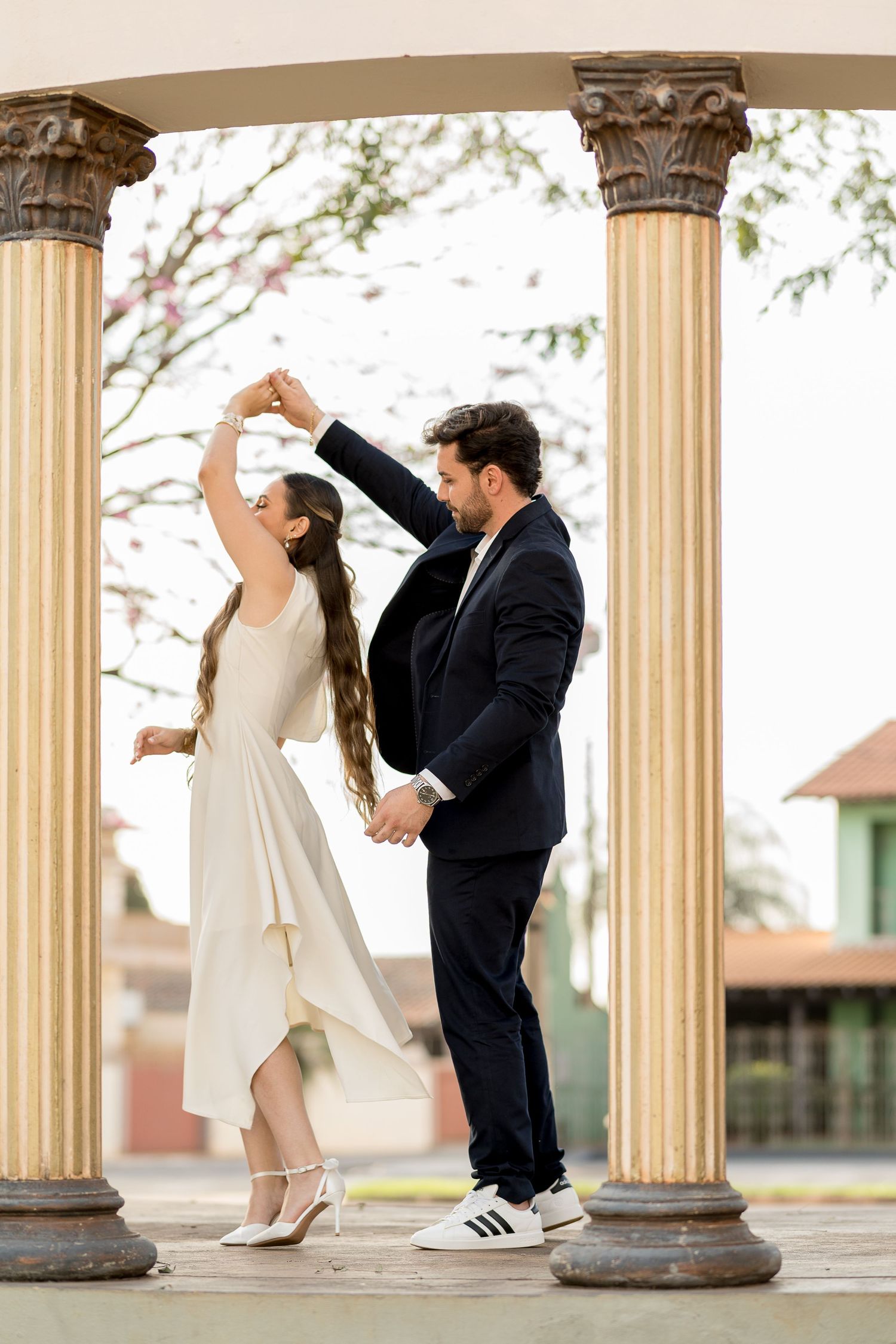Elegant couple dancing between classic columns, woman in white midi dress spinning, man in navy suit and sneakers.